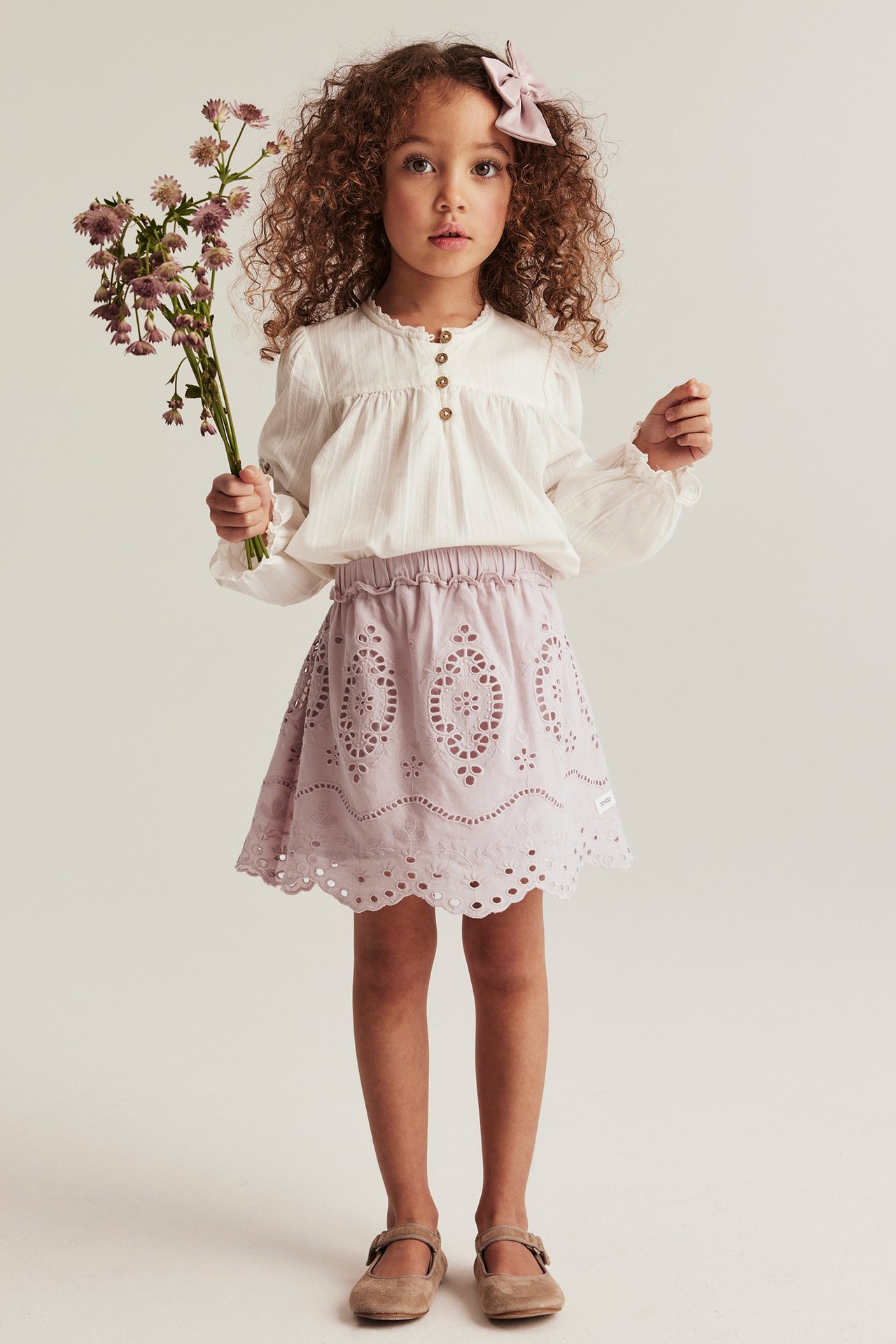 Framifrån: Barn i rosa broderad A-linjekjol med resår i midjan, vit blus, beiga skor, håller blommor.