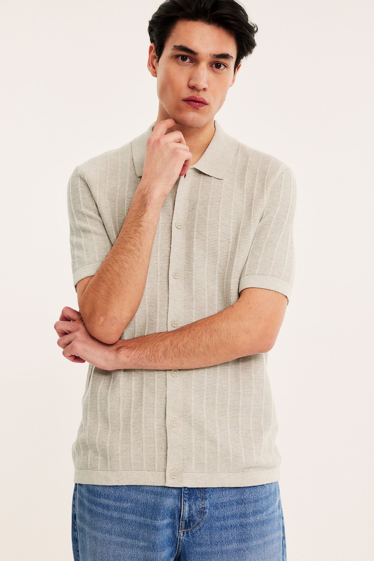 Edestä kuvattu mies beigessä lyhythihaisessa neulotussa pikeepaidassa ja sinisissä farkuissa.