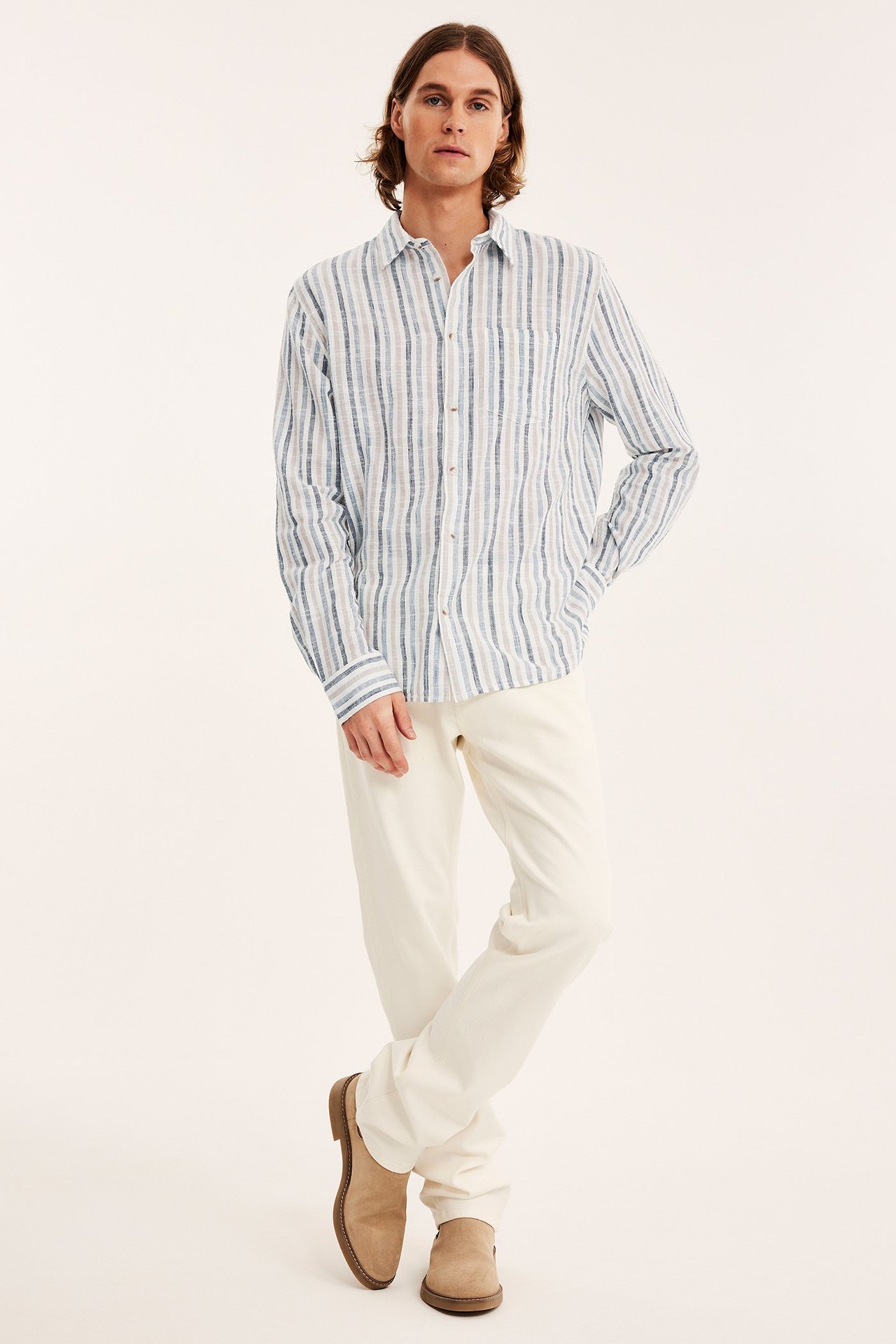 Framifrån: man i ljusblå, vit och beige randig bomullsskjorta, benvita byxor och beiga loafers.