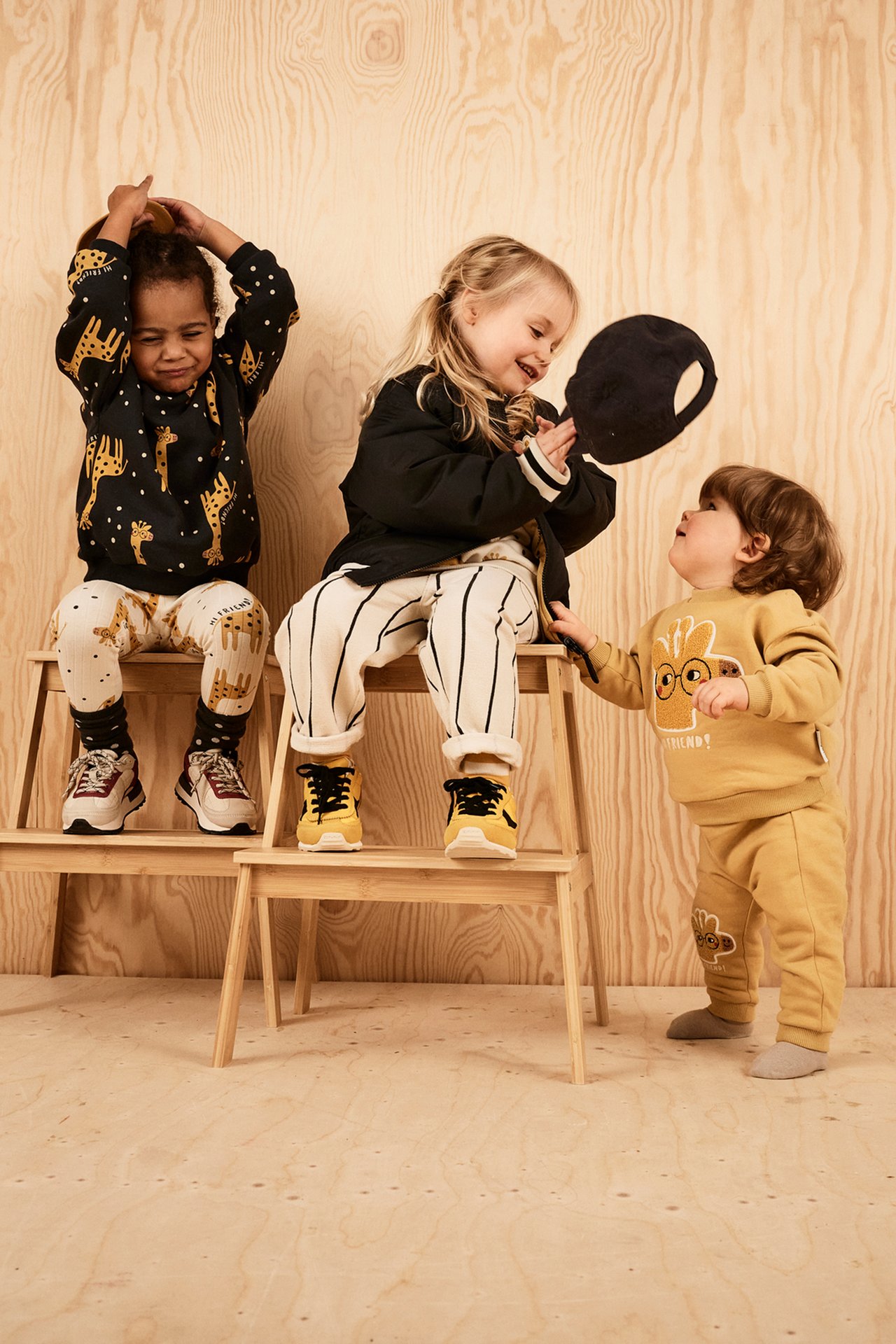 Framsida: Tre barn. Ett bär beiga randiga mjukisbyxor, svart jacka, gula sneakers.