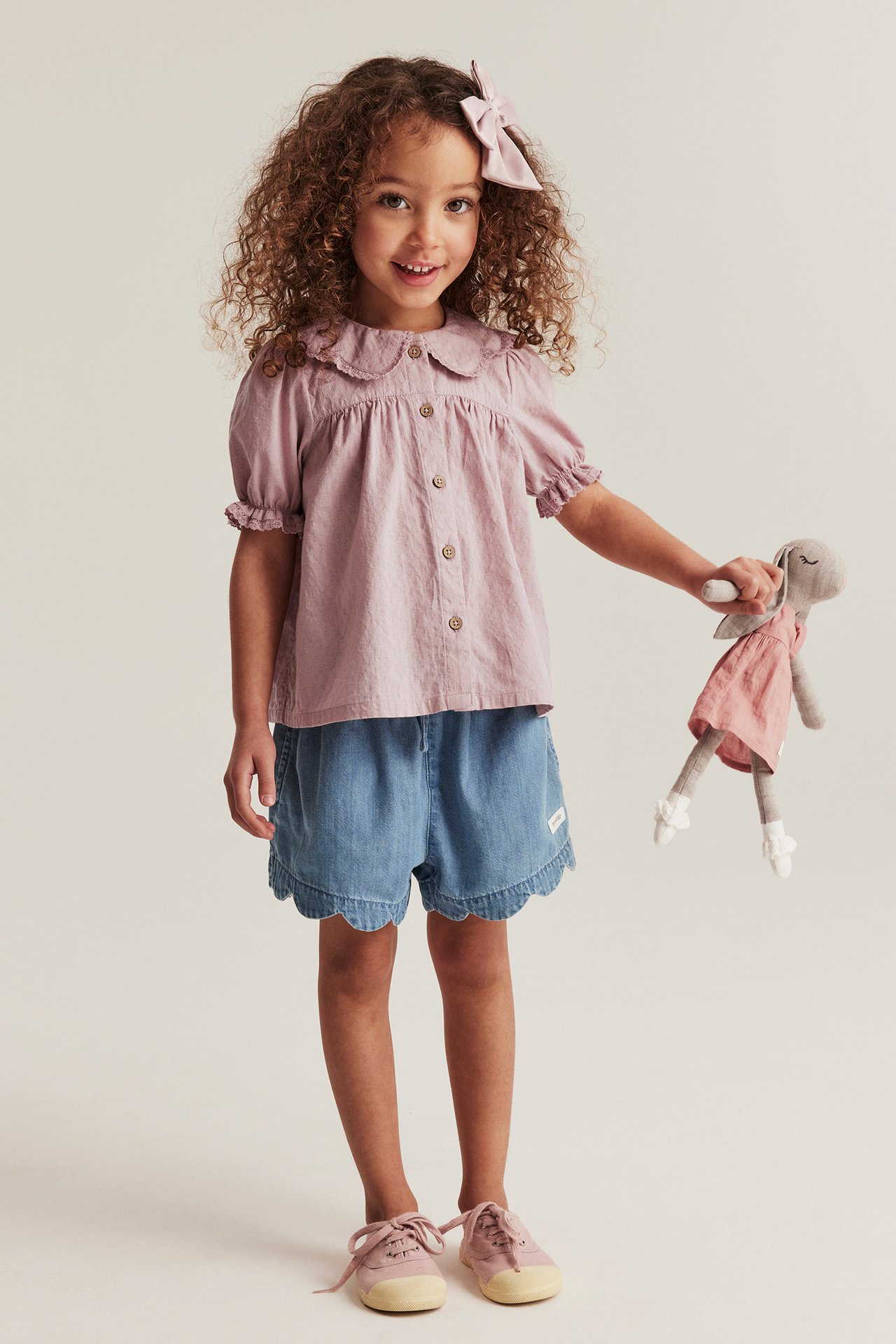 Barn i rosa teksturert bluse, blå shorts, rosa sko, rosa sløyfe, holder en kanindukke.