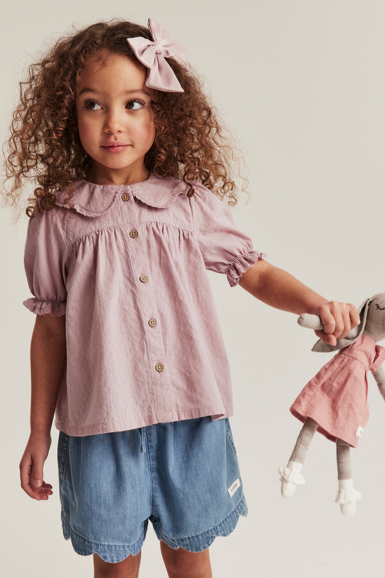 Forfra: Barn i rosa bomullsbluse med blondekrage, blå shorts, rosa sløyfe, holder en kanindukke.