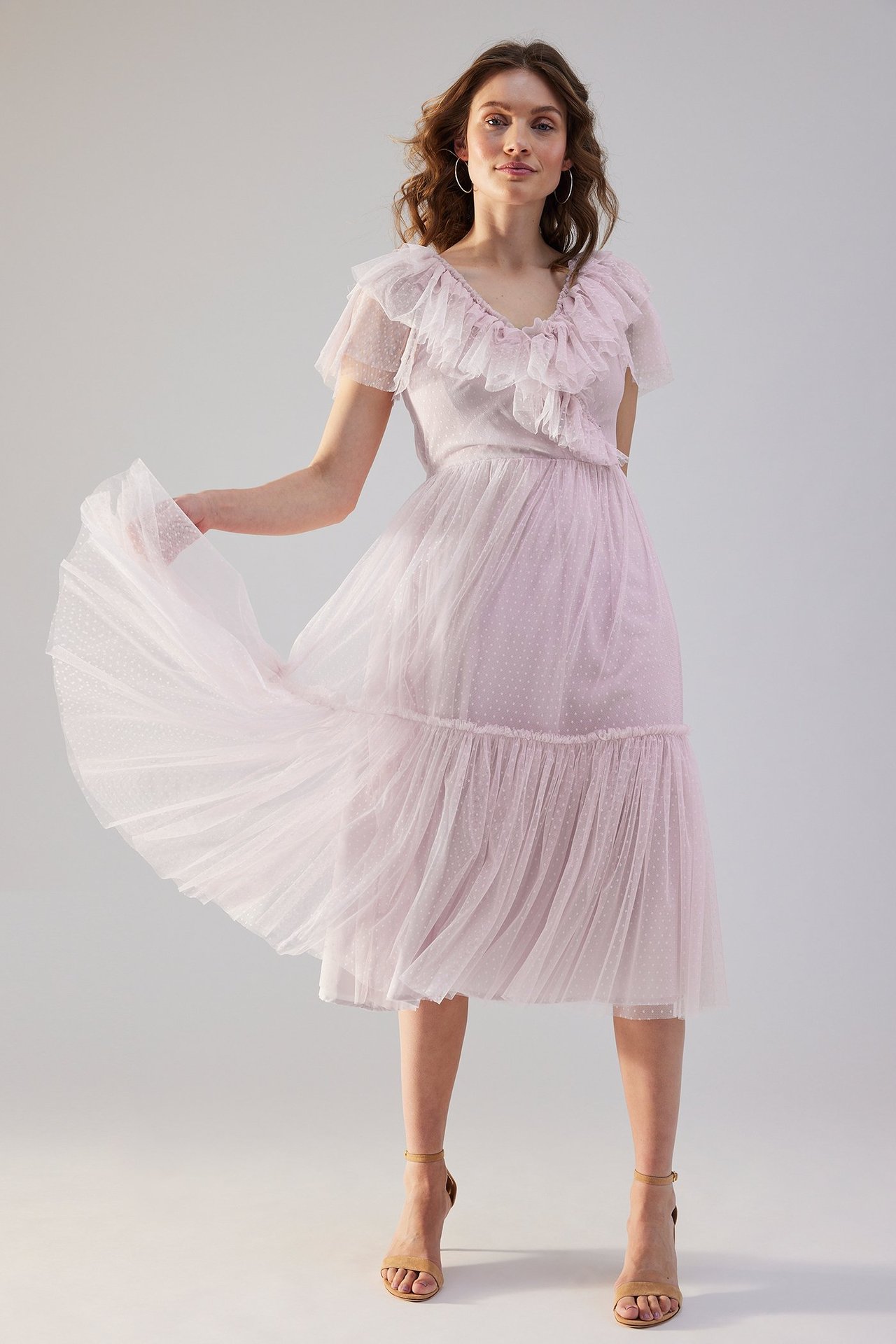 Edestä: nainen yllään vaaleanlila röyhelöinen verkkomekko pilkuilla ja nudenväriset korkokengät.