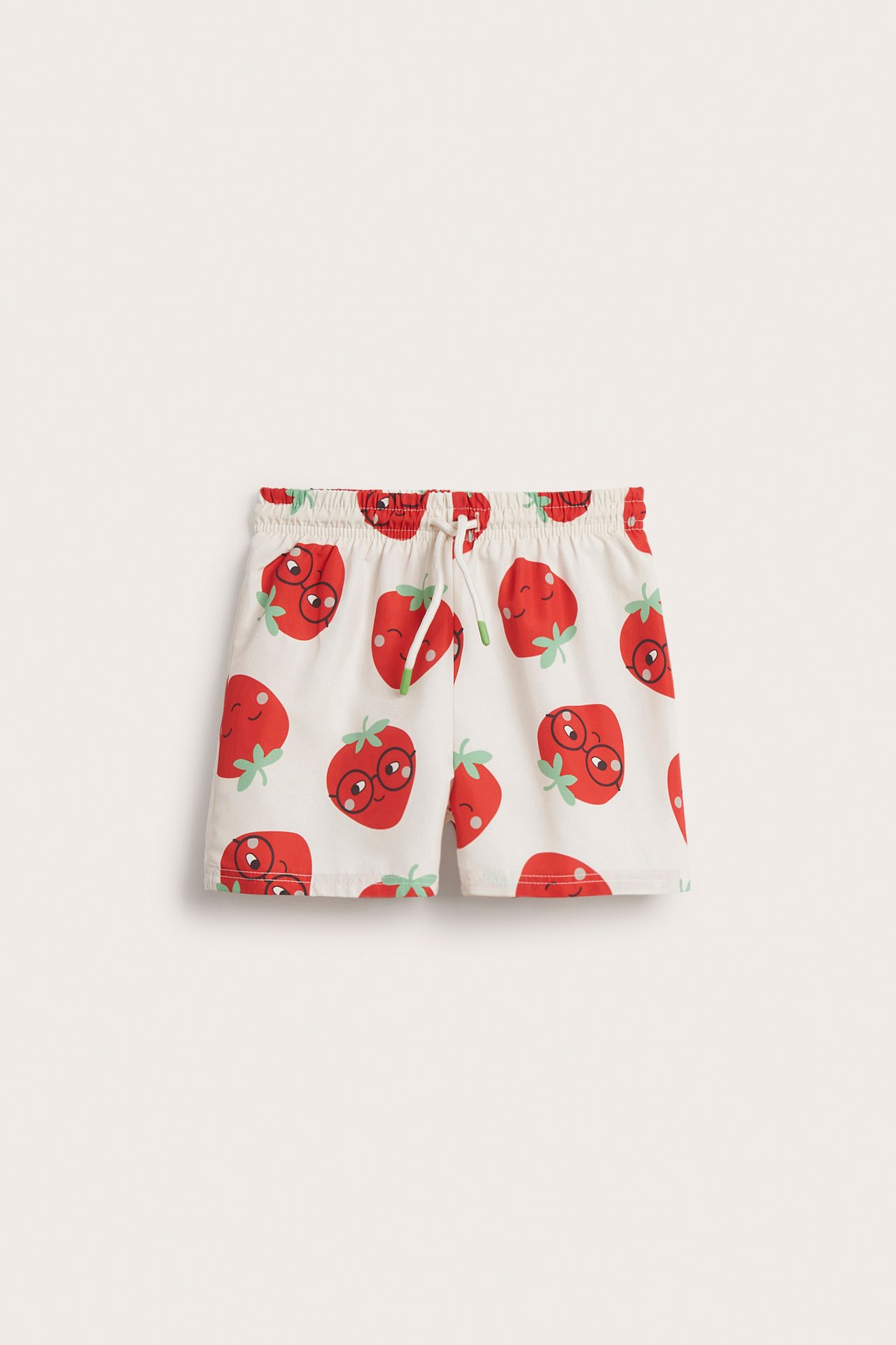 Frontvy av barnens ljusbeige vävda badshorts med rött jordgubbsmönster, resår i midjan och dragsko.
