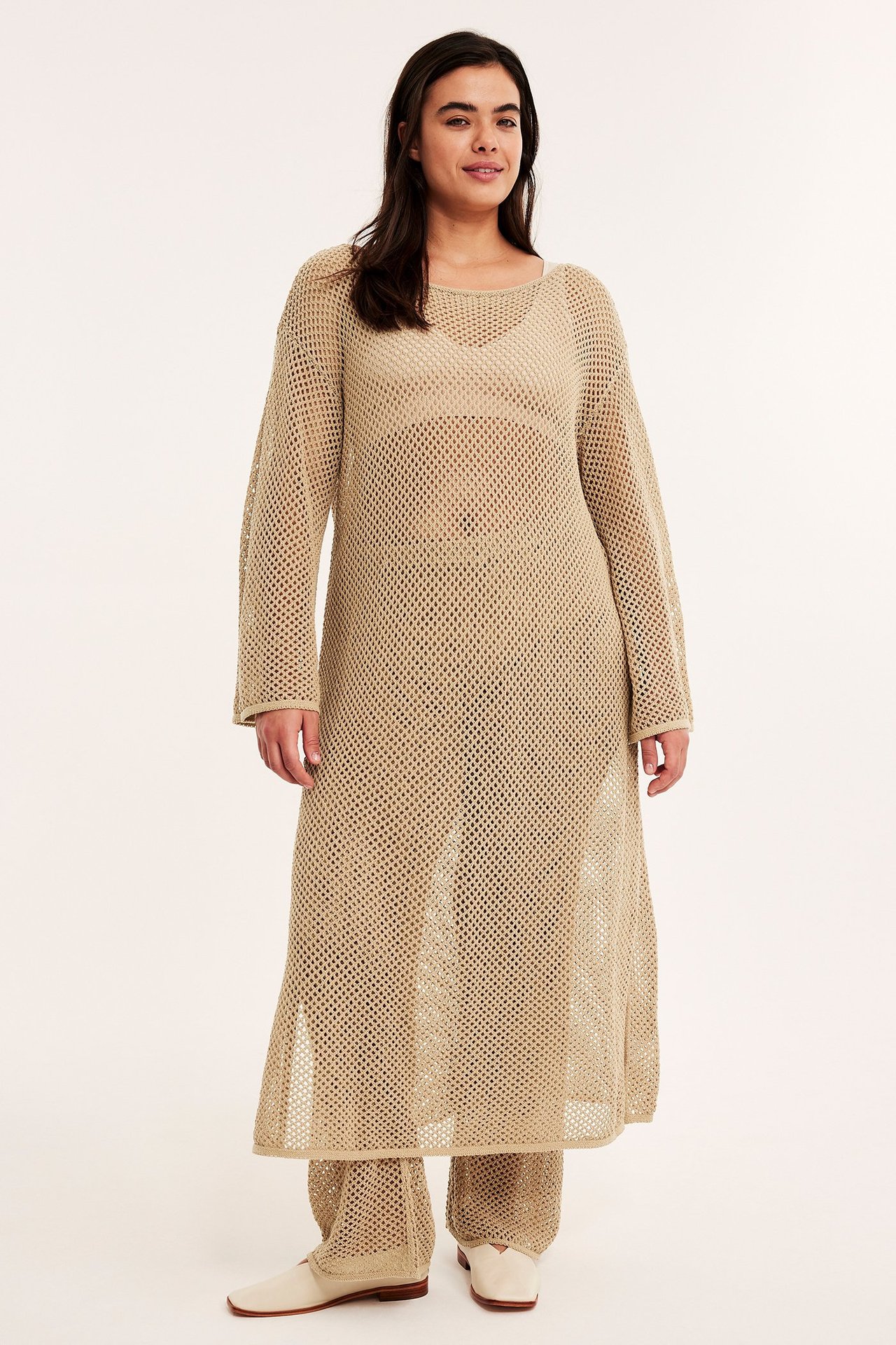 Framifrån: kvinna i mörkbeige hålstickad midiklänning, matchande byxor och ljusa platta skor.