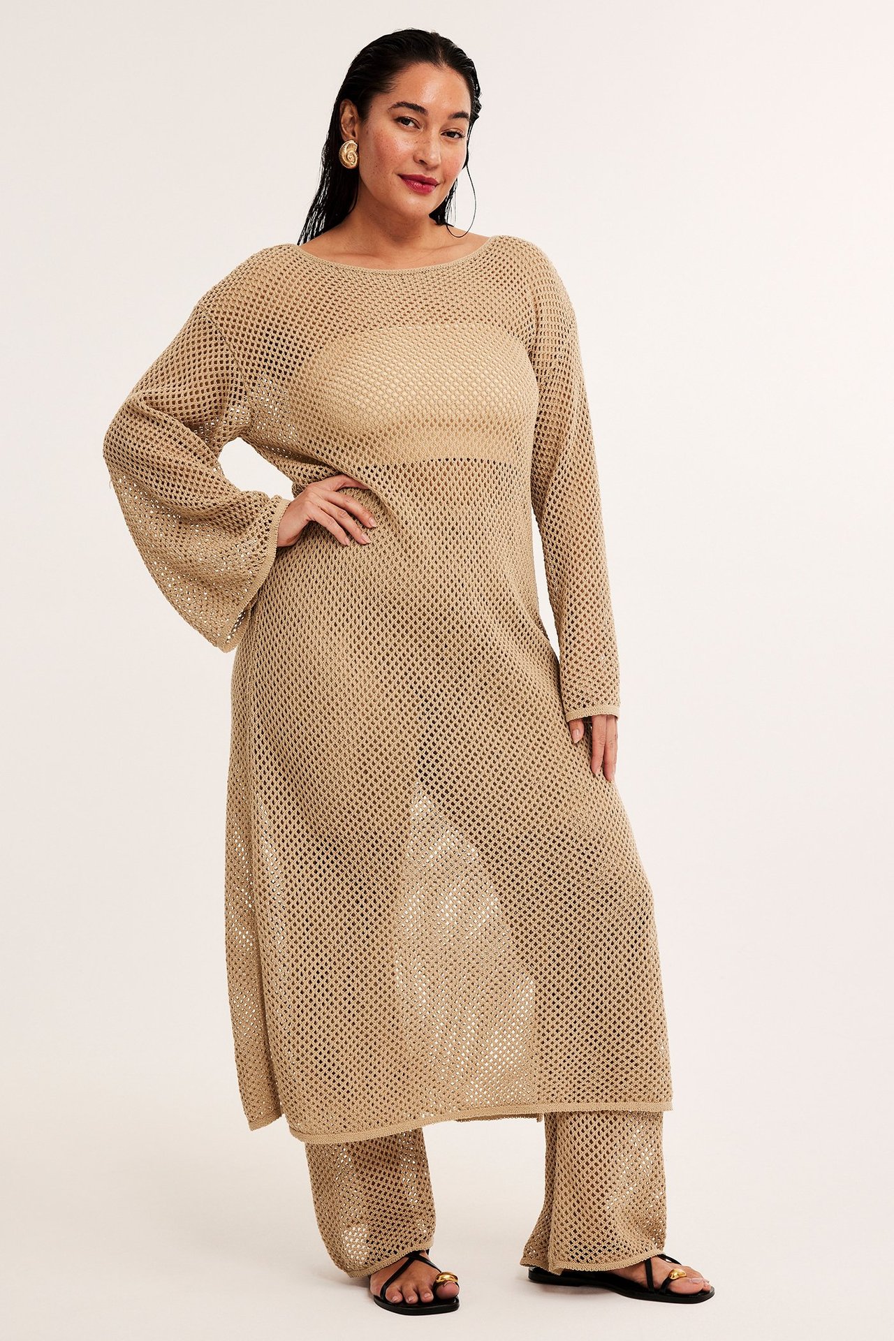 Frontvy av kvinna i lång, lös mörkbeige hålstickad klänning och matchande byxor.