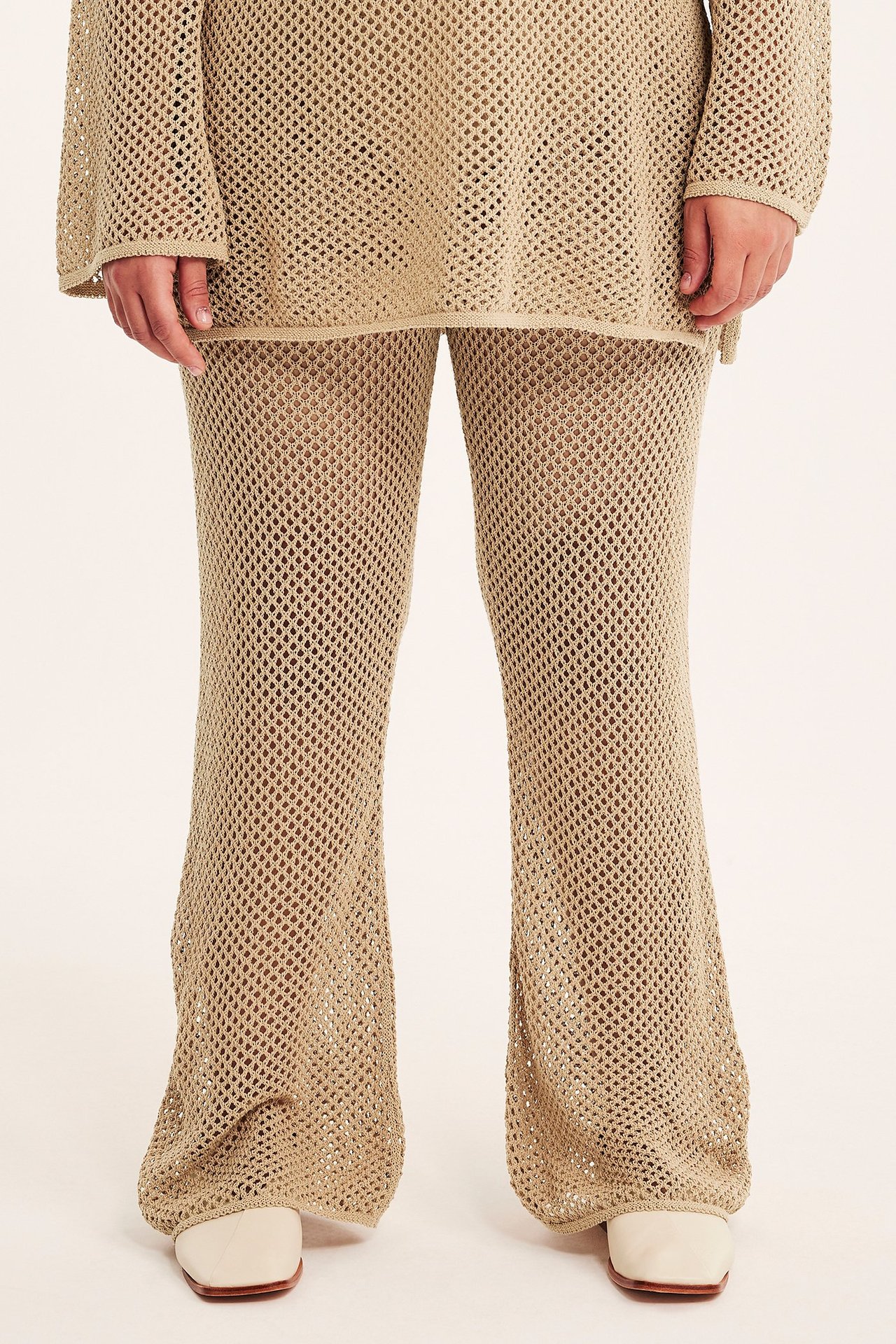 Framifrån: Kvinna bär mörkbeige hålstickade utsvängda byxor, matchande topp och vita skor.
