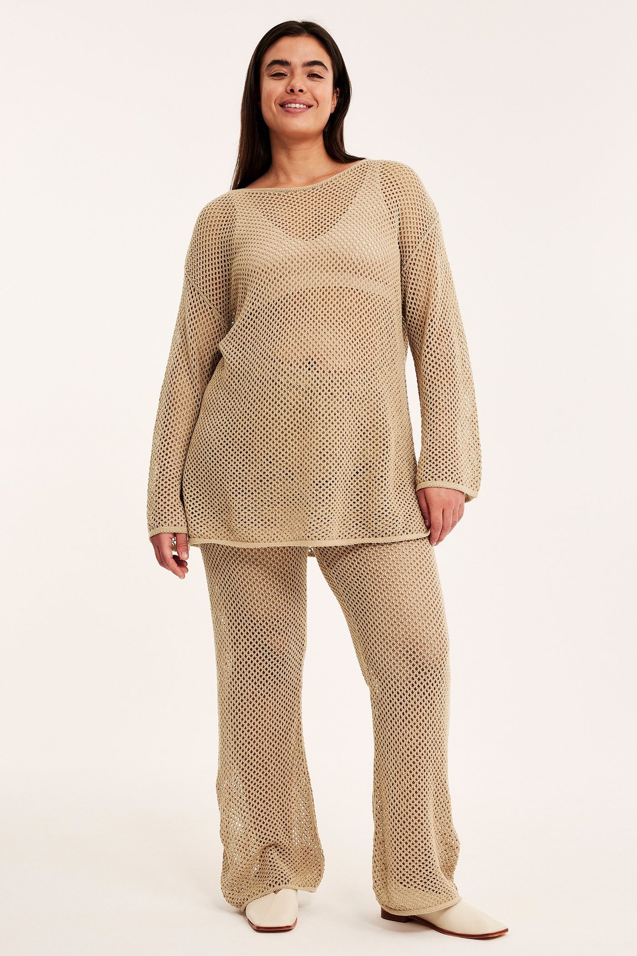 Widok z przodu kobiety w ciemnobeżowych ażurowych spodniach, luźnym topie i jasnobeżowych butach.