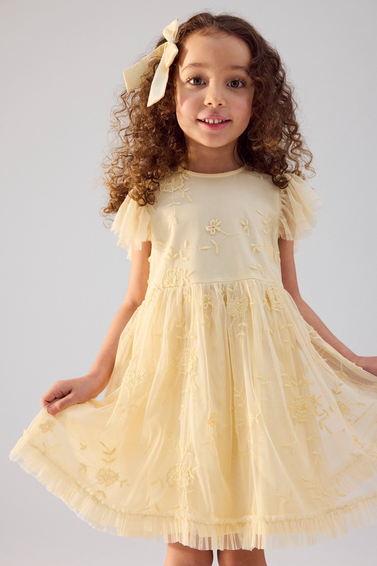 Widok z przodu dziecka w żółtej haftowanej sukience z falbankami i pasującą kokardą we włosach.