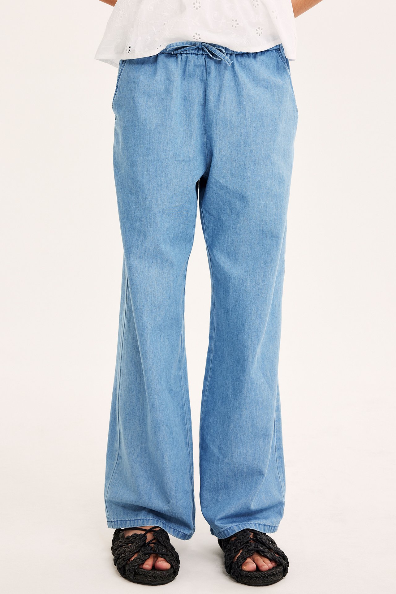Frontvy av ett barn i ljusblå denimbyxor med dragsko, en vit topp och svarta flätade sandaler.