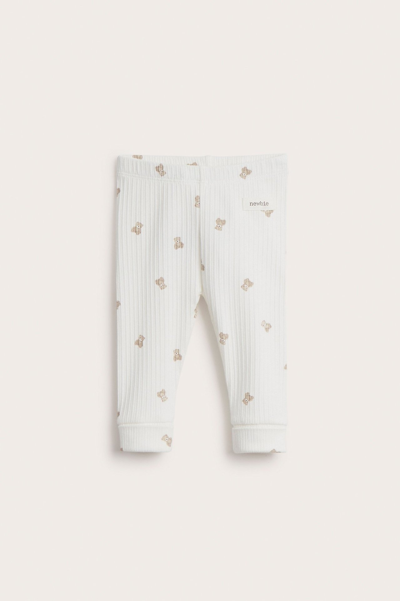 Framifrån: Offwhite ribbade baby leggings med brunt nallemönster och 'newbie' etikett.