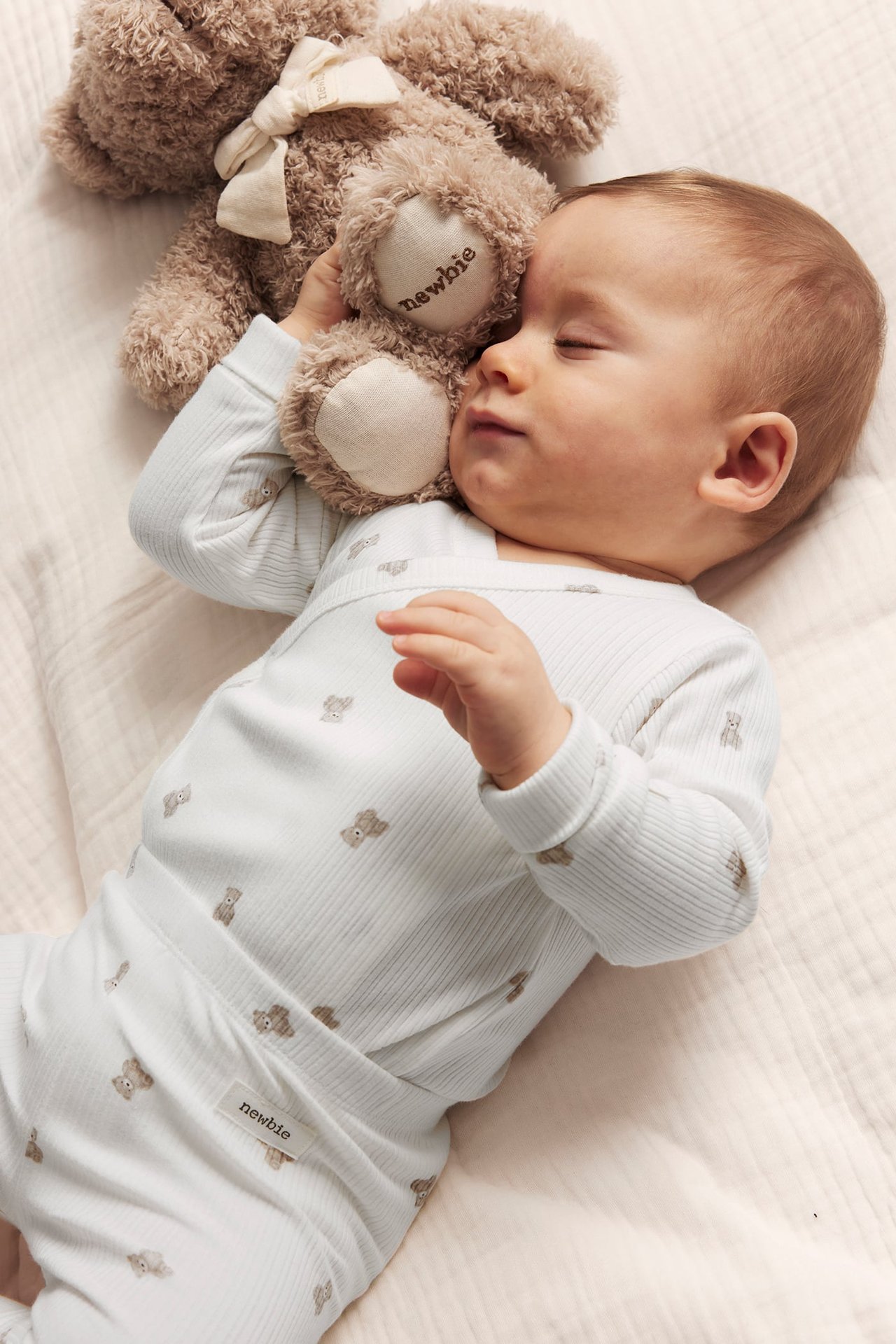 Sovende baby i offwhite ribbet omslagsbody med bamsemønster, holder en brun kosebamse.