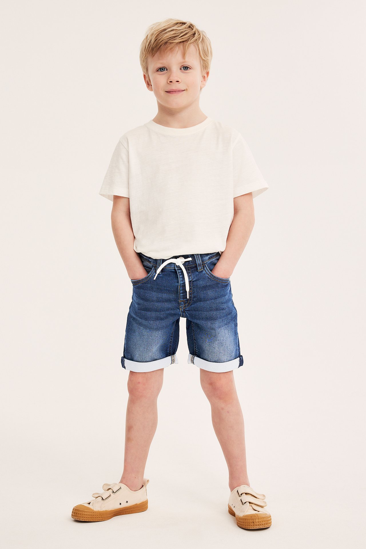 Edestä: poika sinisissä denim-jogger-shortseissa valkoisella nyörillä, valkoisessa t-paidassa ja beigessä lenkkareissa.