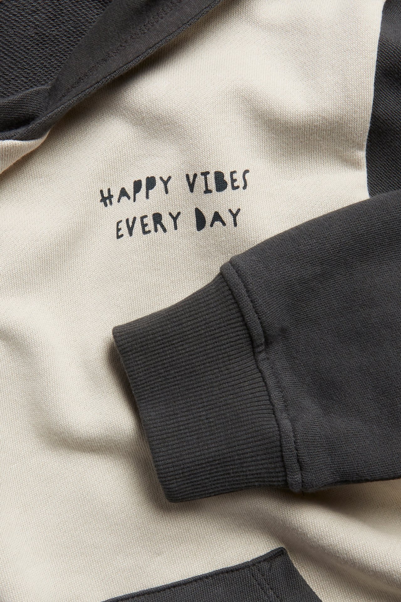 Zbliżenie na dziecięcą bluzę bawełnianą w beżowo-ciemnoszare bloki z napisem "HAPPY VIBES EVERY DAY".