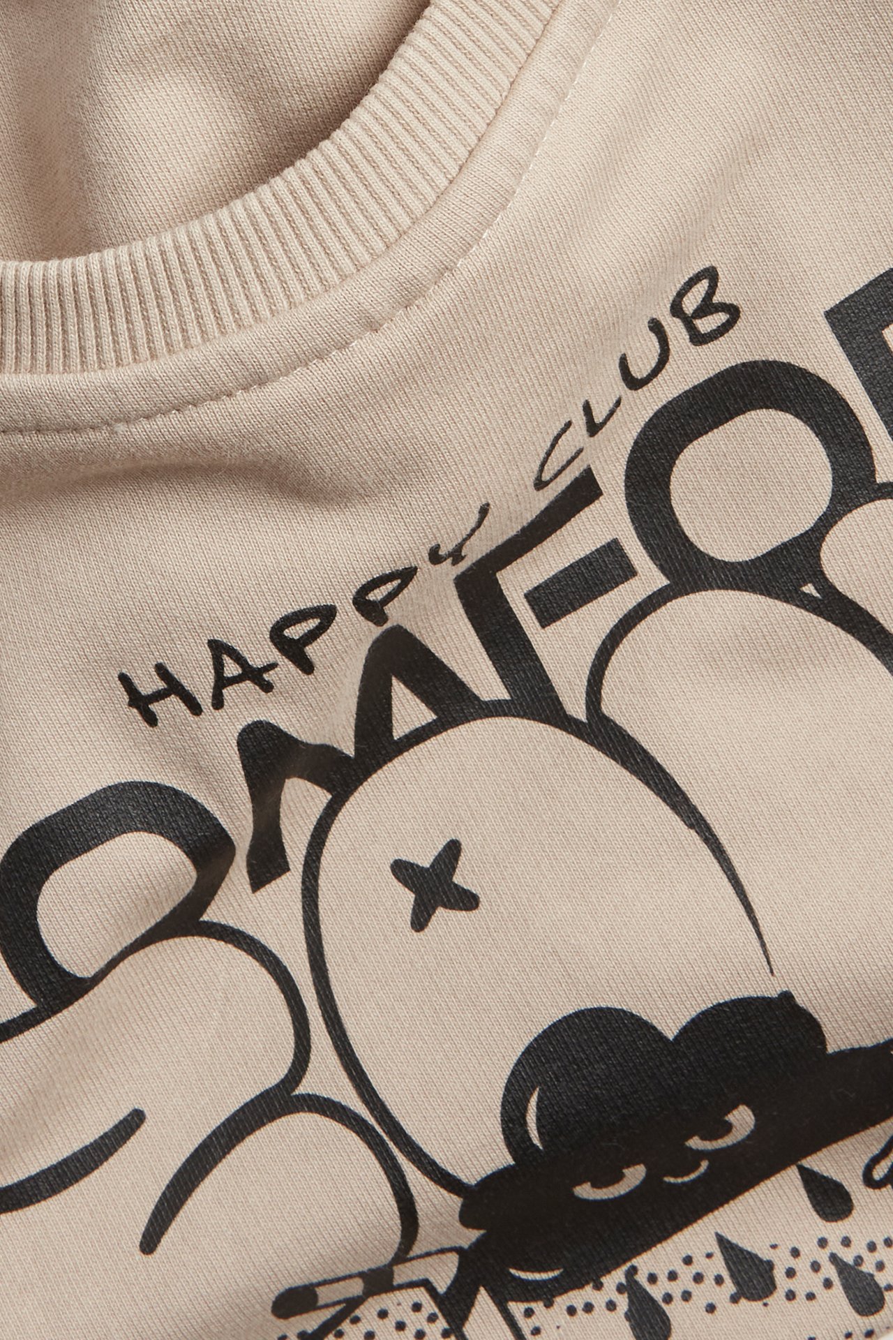 Närbild på en beige bomullssweatshirt för barn med svart 'HAPPY CLUB' grafiskt tryck.