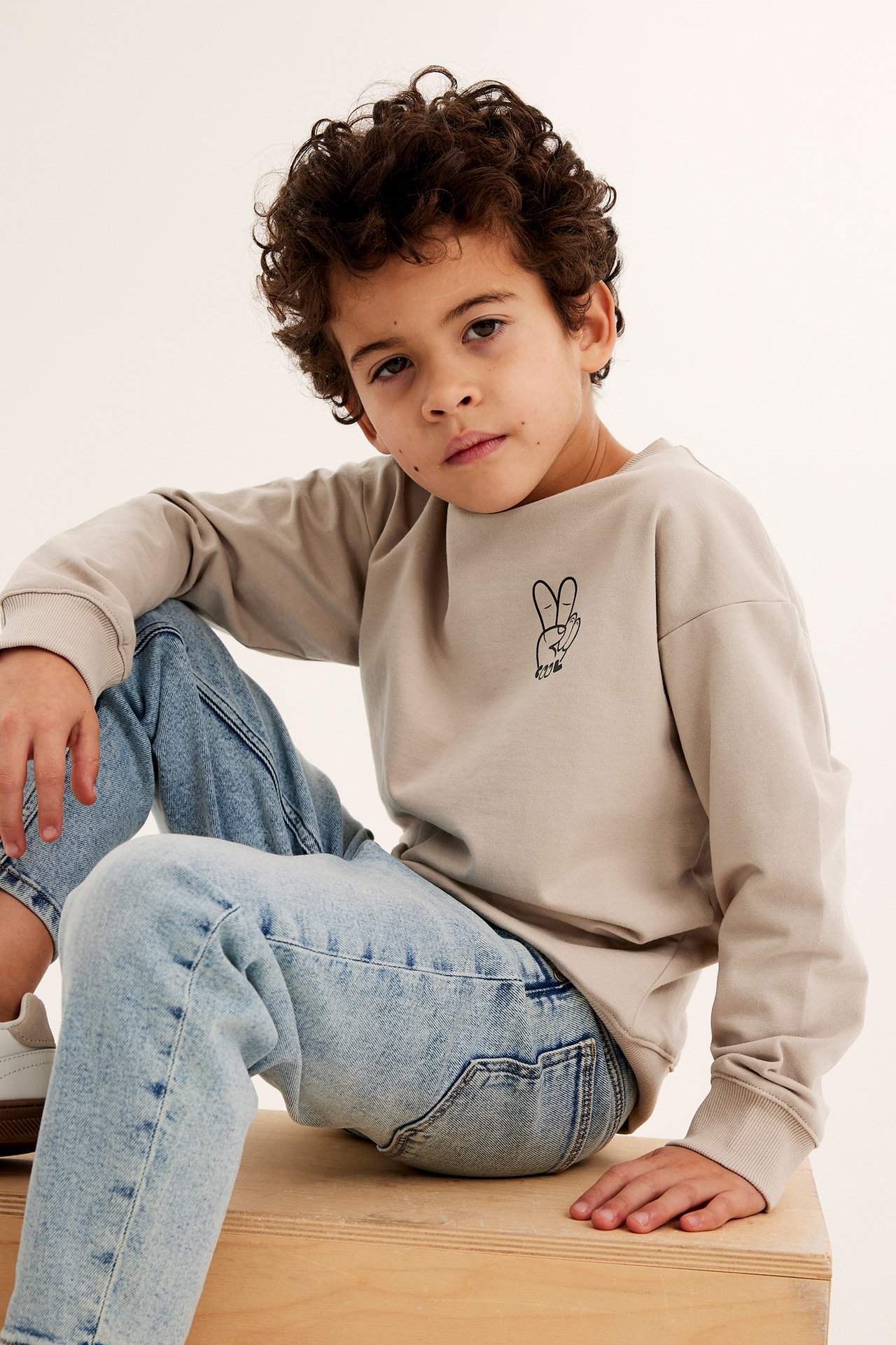 Framifrån, ett barn i beige bomullssweatshirt med svart fredstecken-tryck och ljusblå jeans.