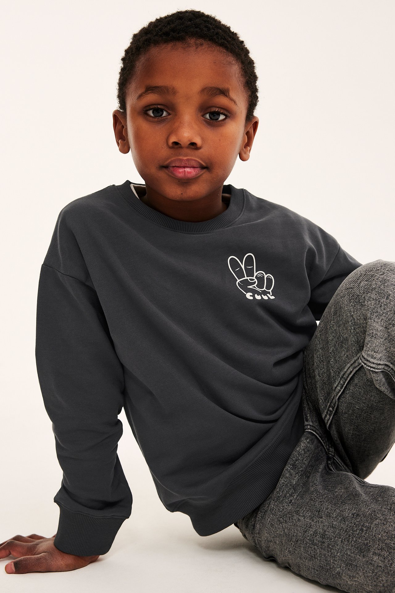 Framifrån: Kid i mörkgrå bomullssweatshirt med vitt fredstecken-tryck och mörkgrå jeans.