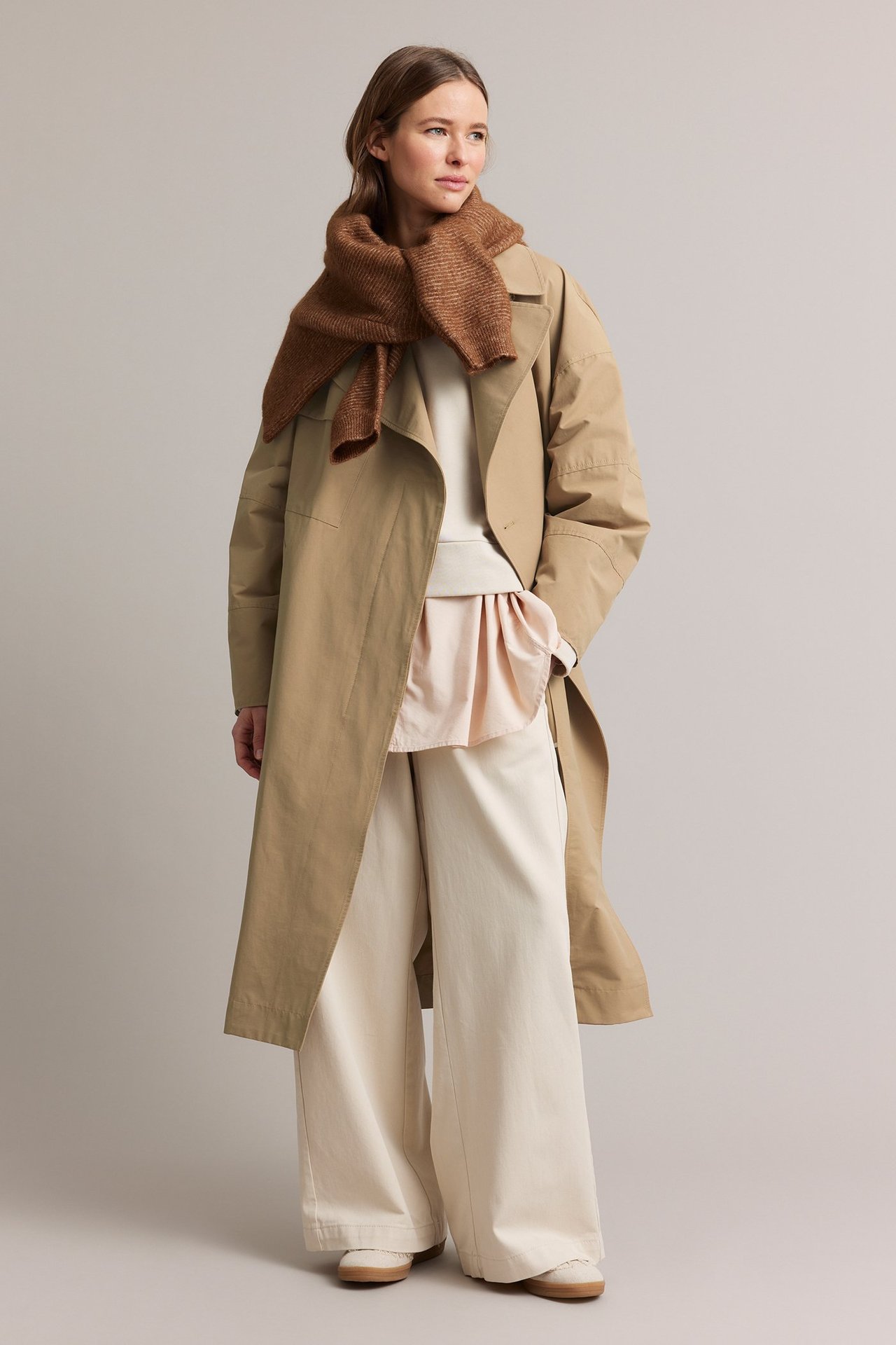Frontvy: Kvinna i beige trenchcoat, brun stickad halsduk, krämvit topp och vida krämvita byxor.