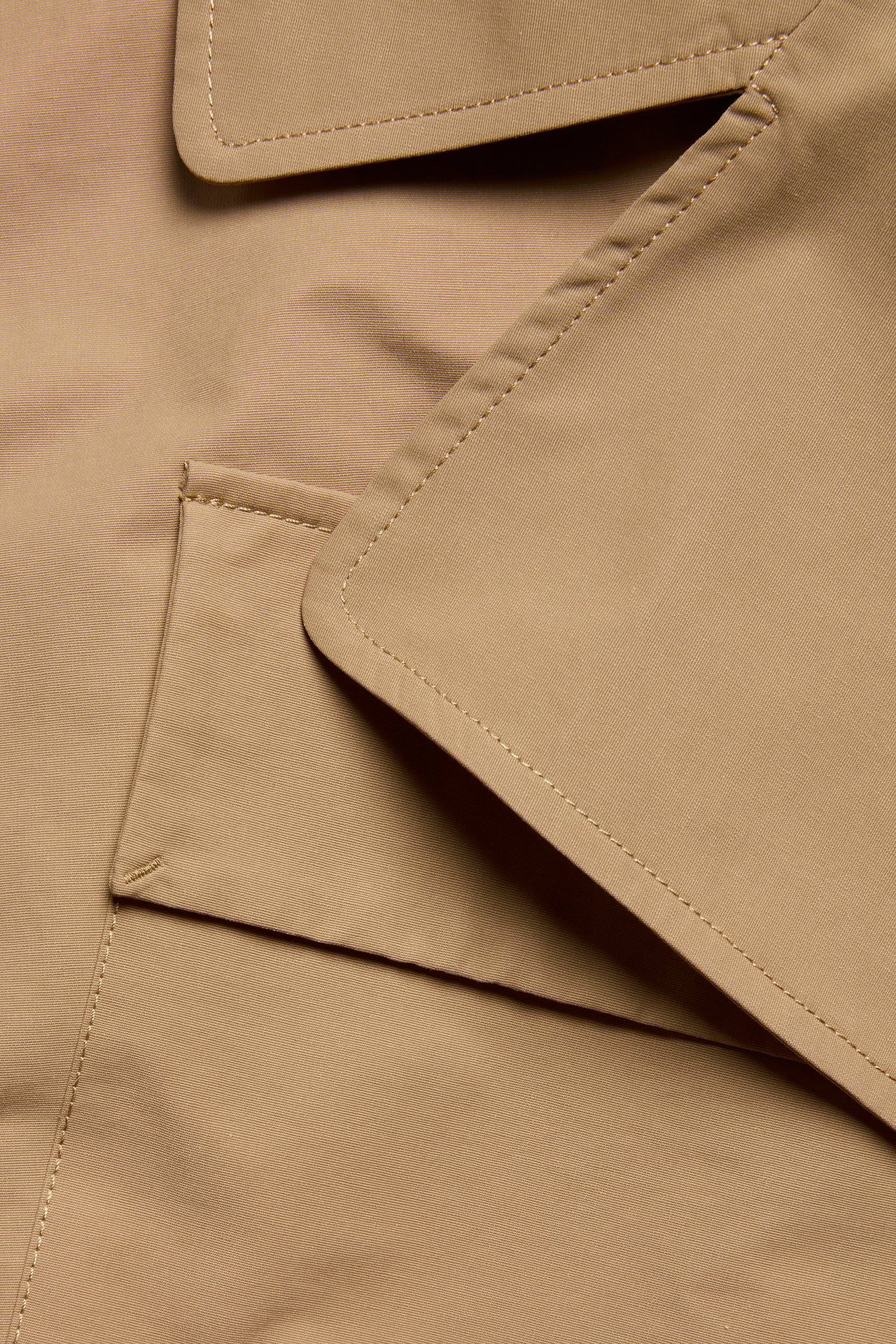 Zbliżenie beżowego płaszcza trenczowego z mieszanki bawełny z widocznymi szwami i klapą kieszeni.