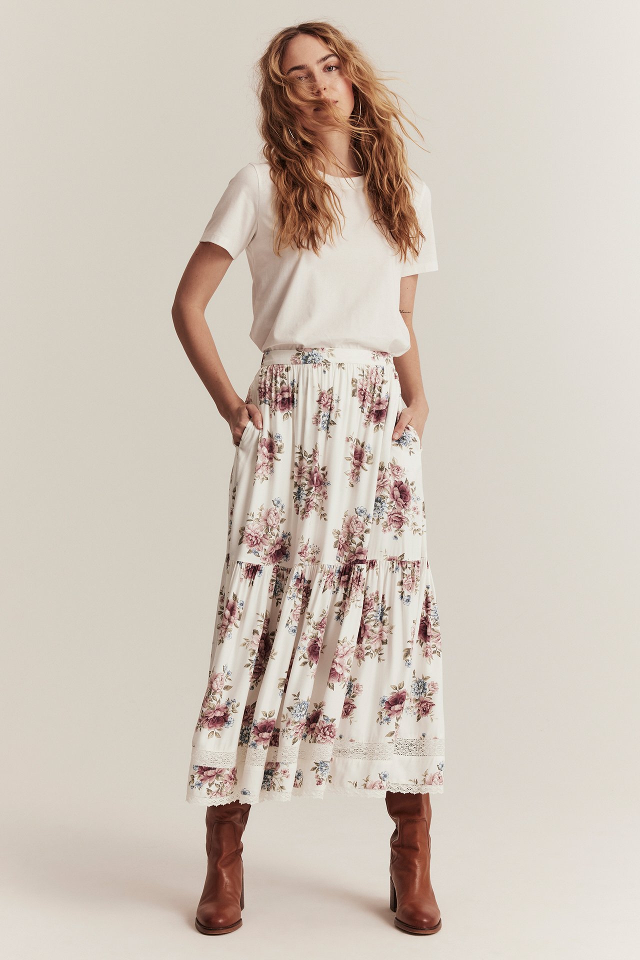 Framifrån: kvinna i offwhite blommig viskos maxikjol med spets, vit t-shirt och bruna stövlar.
