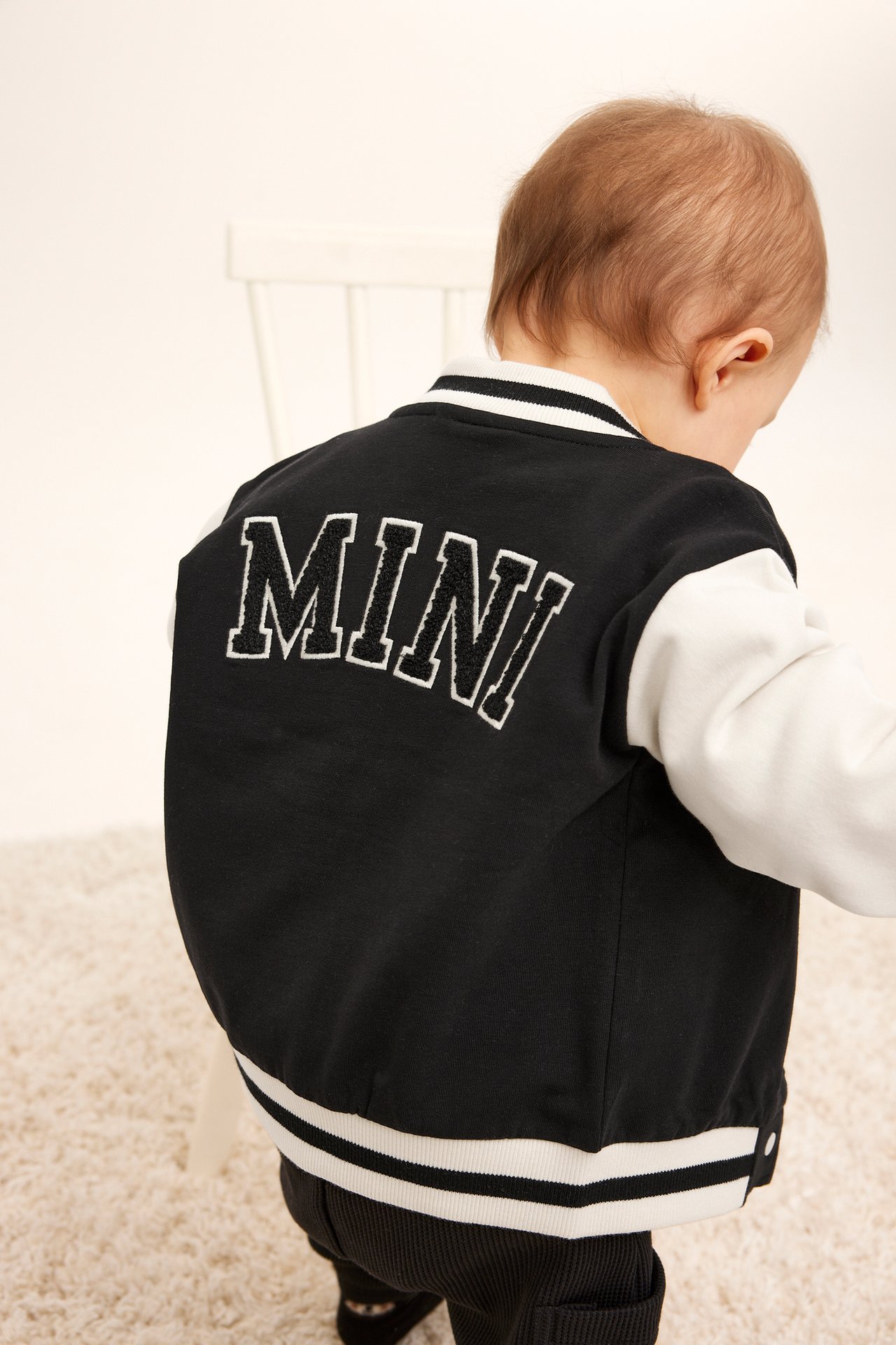Takaa näkymä vauvasta mustavalkoisessa baseball-takissa, jossa "MINI" applikointi ja tummat ribbihousut.