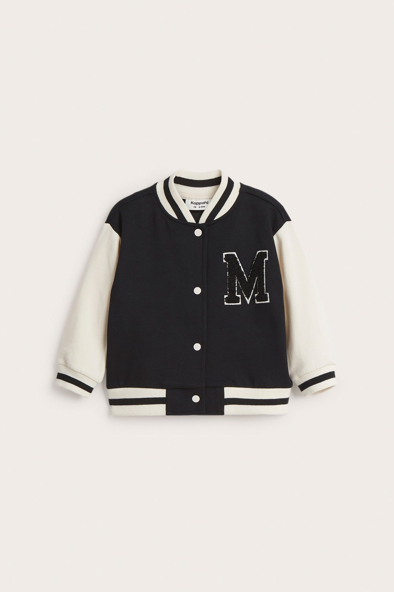 Edestä: lasten musta-kermanvärinen baseball-takki valkoisilla napeilla ja 'M'-froteeapplikaatiolla.