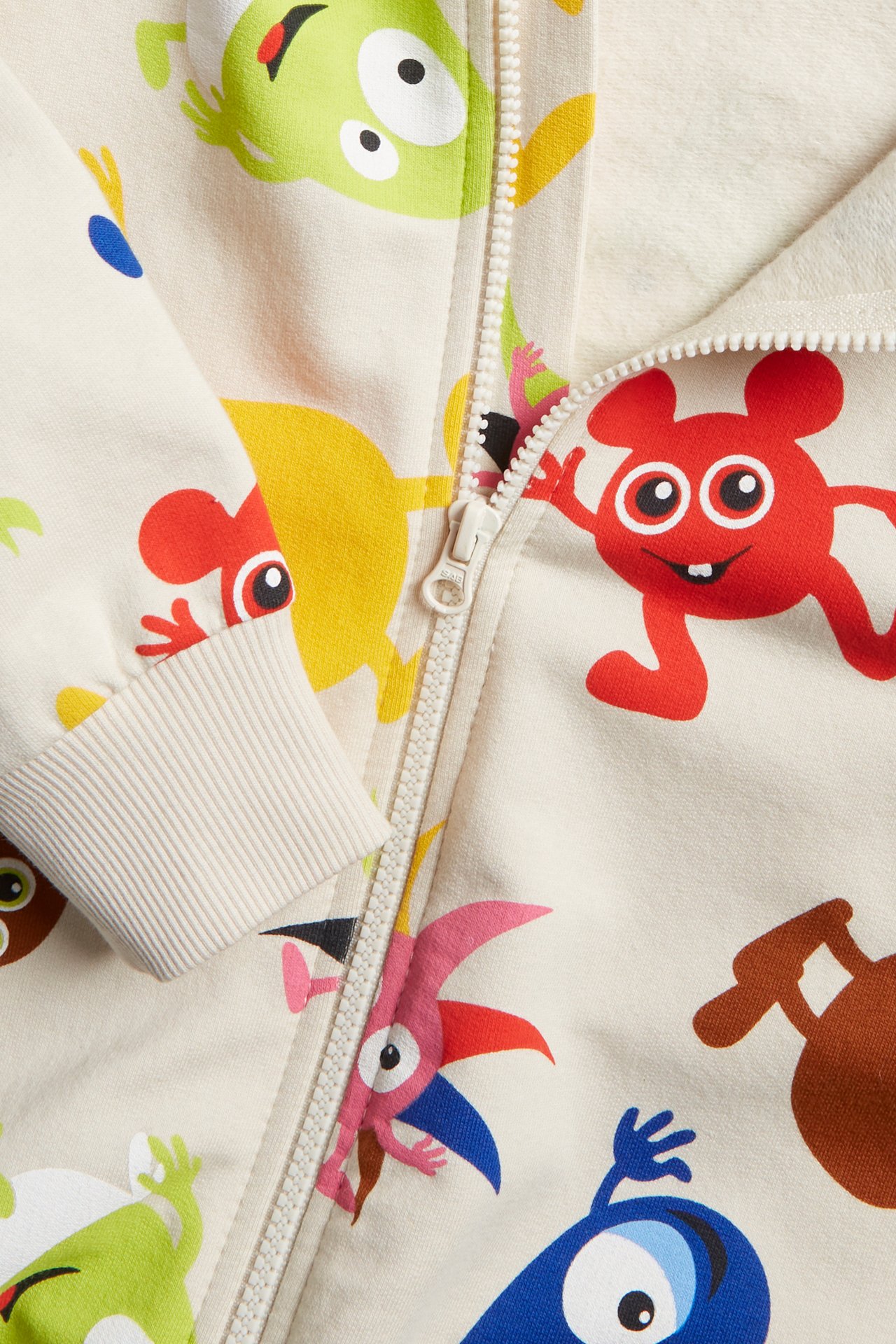 Närbild på ljusbeige babyjumpsuit med färgglatt Babblarna-mönster, dragkedja fram och ribbade muddar.