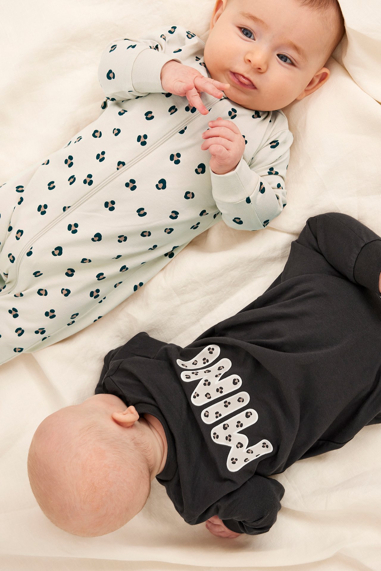Två bebisar bär långärmade pyjamasar: en beige leopardmönstrad med dragkedja, en mörkgrå med 'MINI'-tryck.