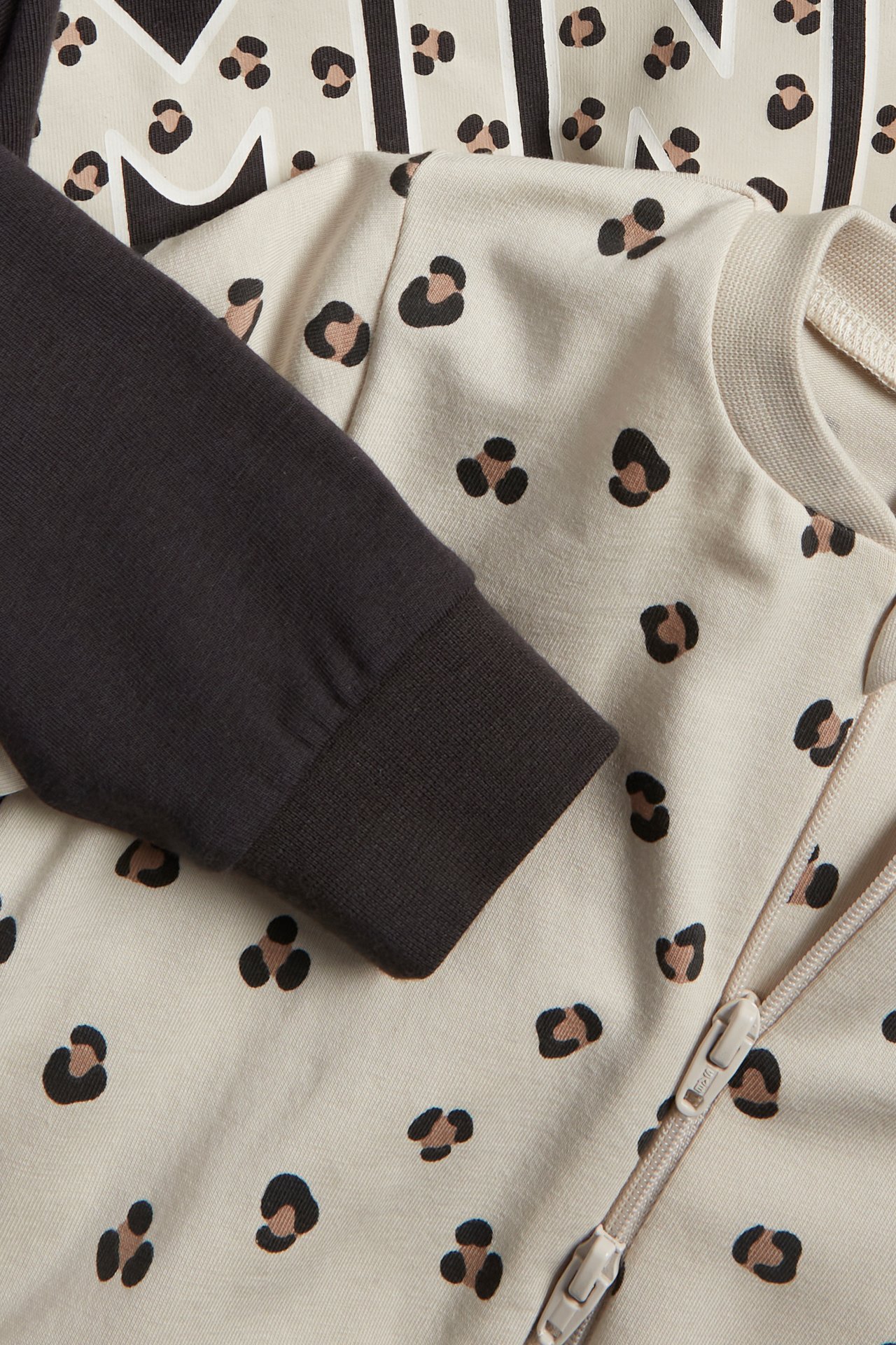 Närbild: Beige leopardmönstrad baby-romper med tvåvägsdragkedja, bredvid en mörkgrå romper med beige tryck.