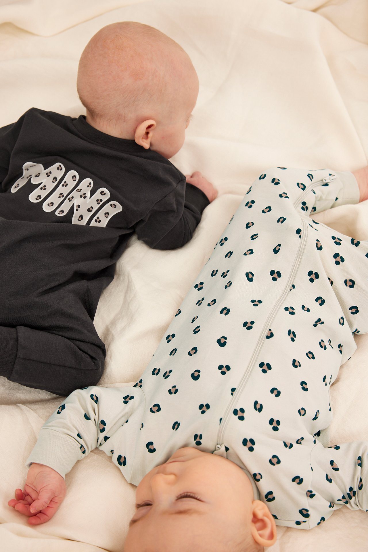Framifrån, en baby i beige leopardmönstrad långärmad pyjamas och en baby i mörkgrå pyjamas.