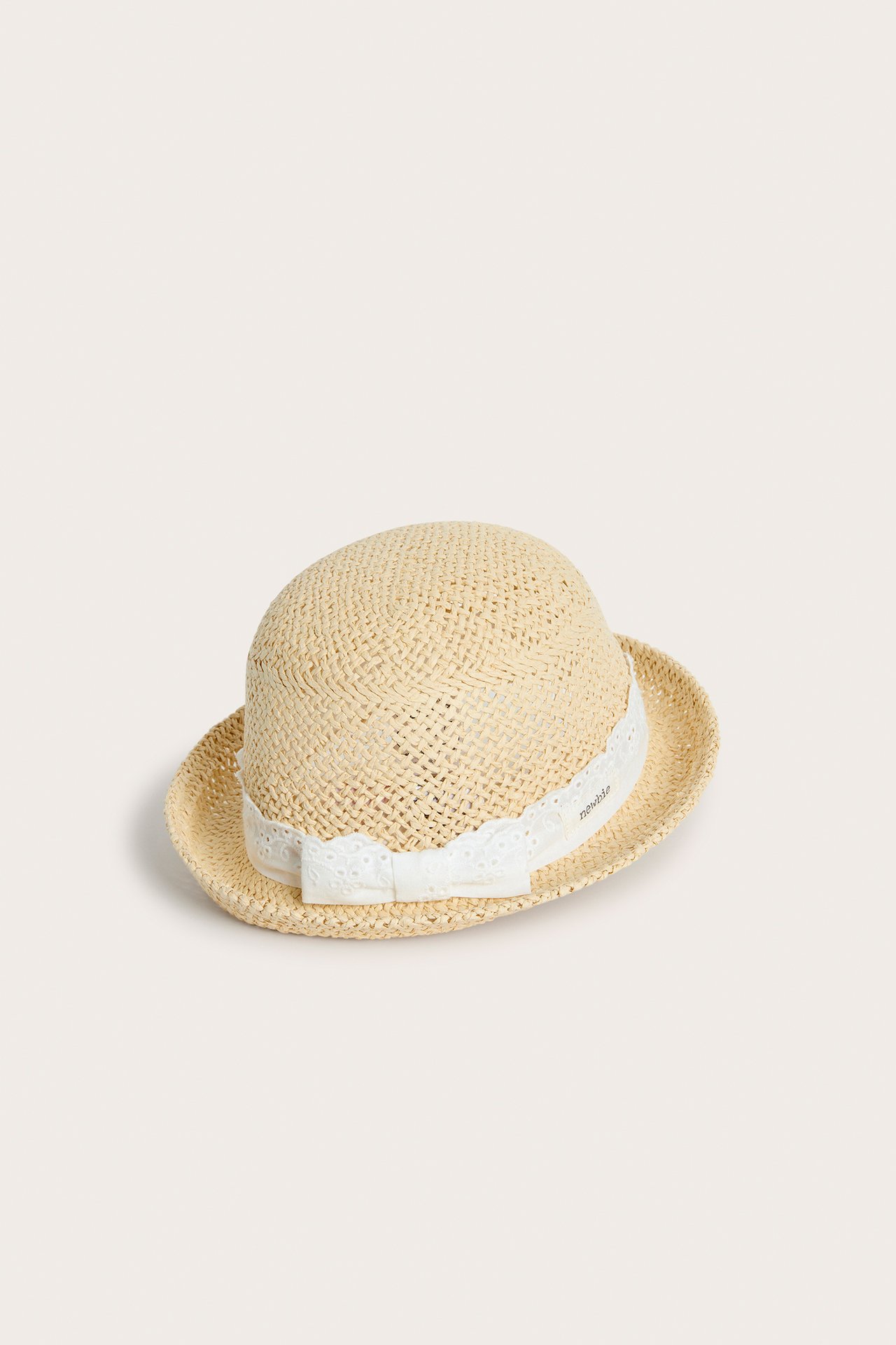 Skrå frontvisning av en lys beige stråhatt for baby/barn med hvitt blondebånd og sløyfe.
