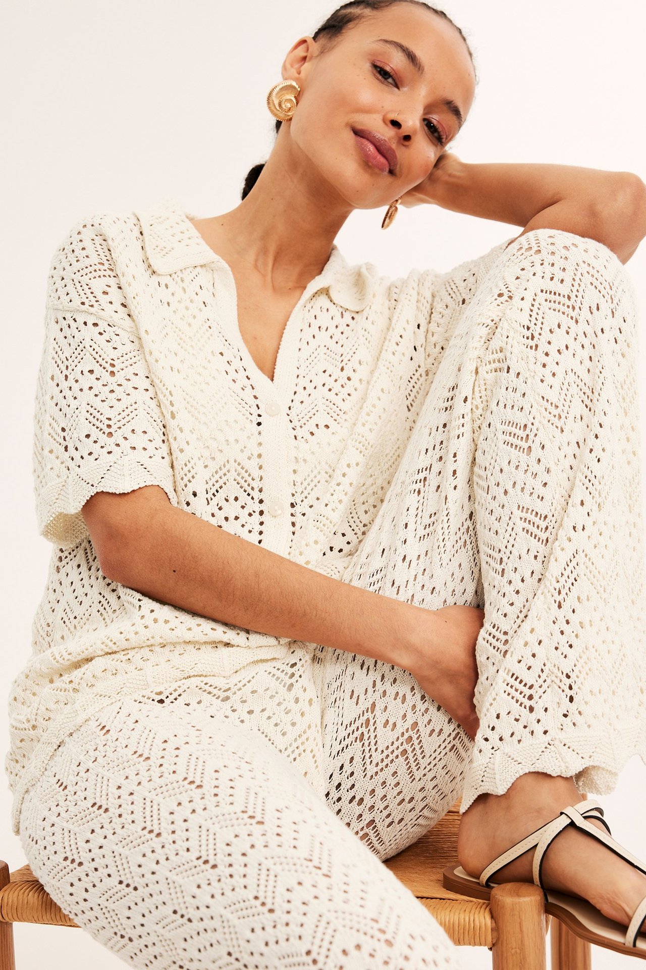 Edestä: nainen yllään vaaleanbeige avoneuleinen lyhythihainen neuletakki, yhteensopivat housut ja sandaalit.