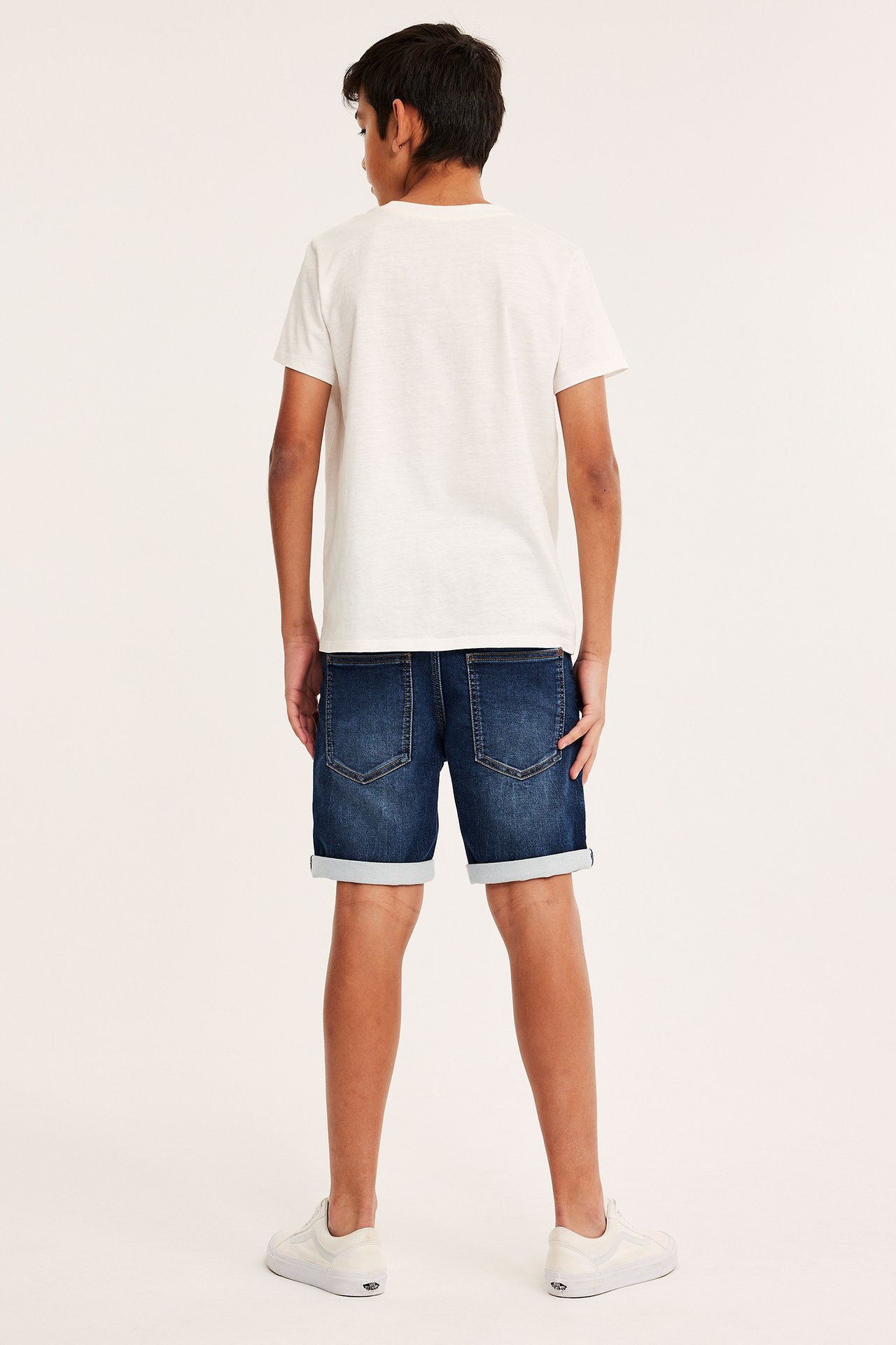 Bakifrån, ett barn i mörkblå jogger-jeansshorts med uppvikta benslut, en vit t-shirt och vita sneakers.