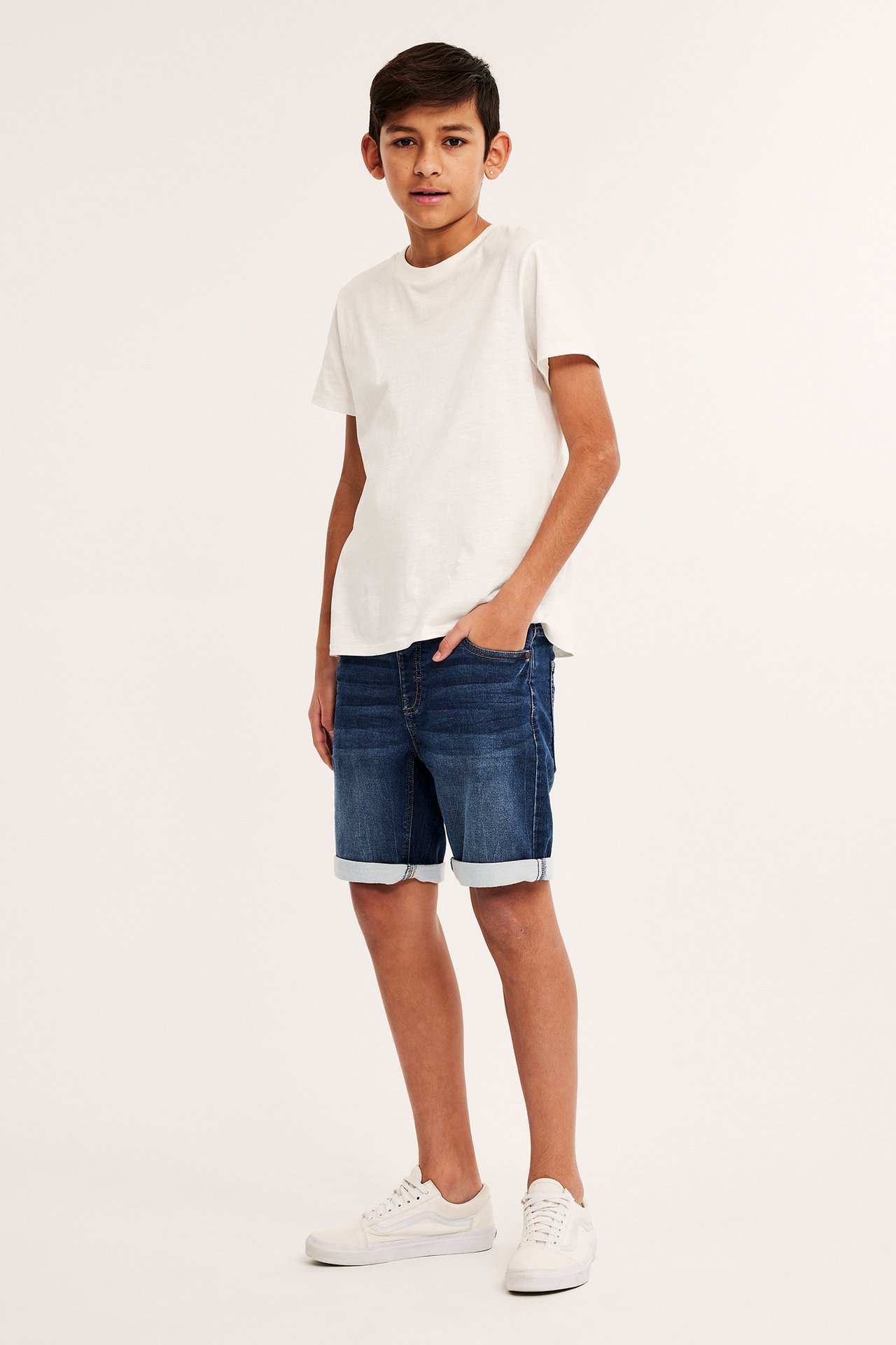 Frontvy av ett barn i mörkblå jogger denimshorts med uppvikta muddar, en vit t-shirt och vita sneakers.