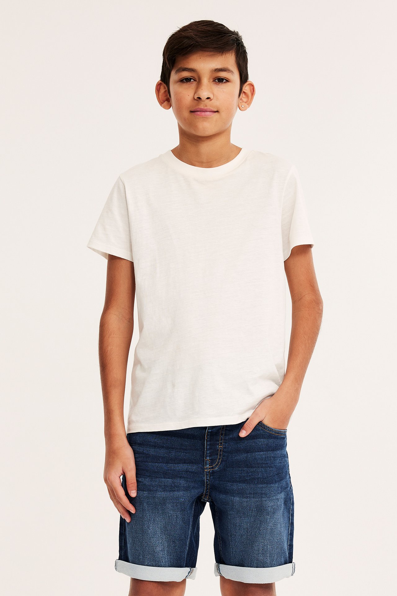 Frontvy av ett barn i mörkblå joggerjeansshorts med uppvikta benslut och en enfärgad krämfärgad t-shirt.