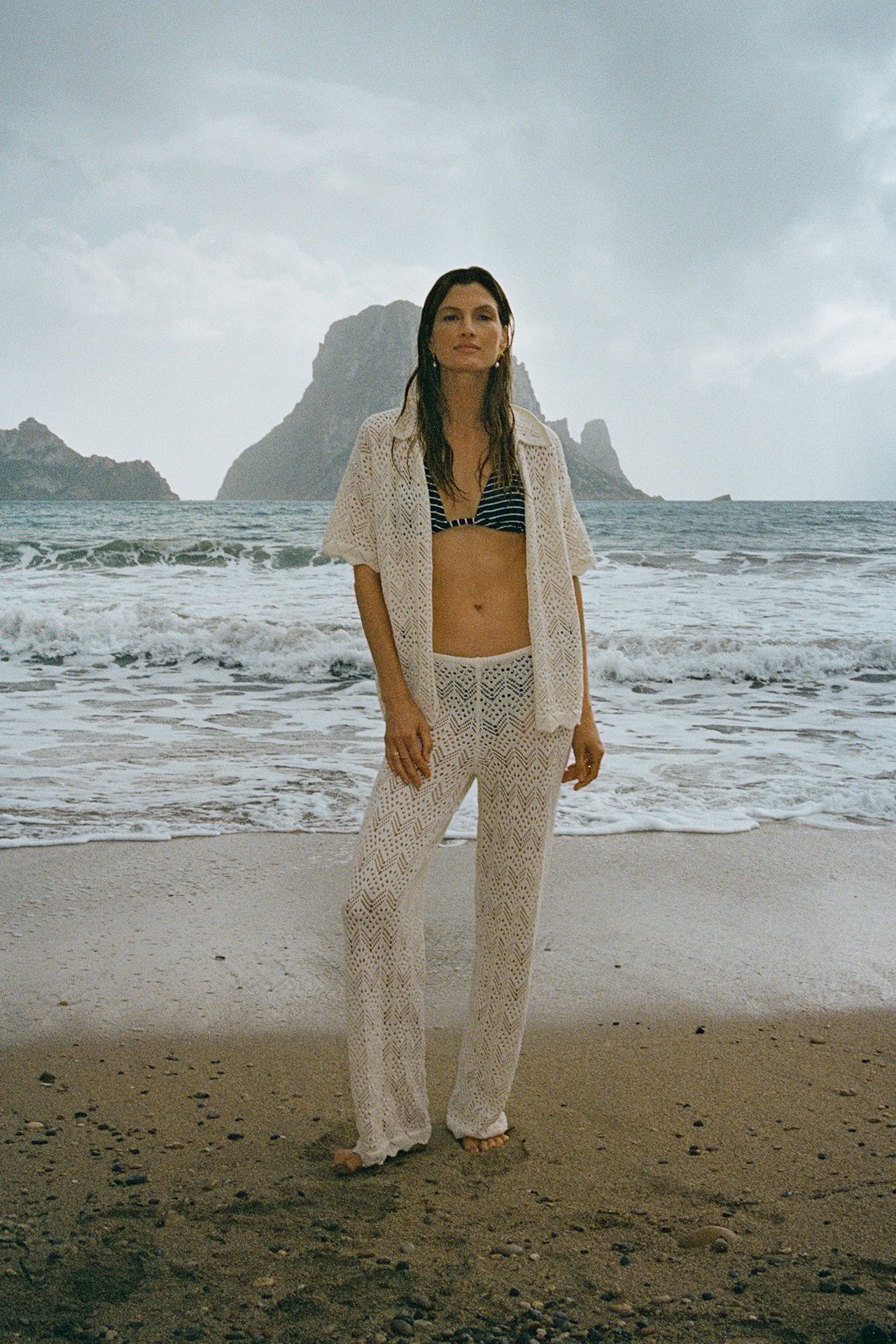 Edestä: Nainen vaaleanbeigeissä neulotuissa virkatuissa leveälahkeisissa housuissa, avoimessa paidassa ja raidallisessa bikinissä.