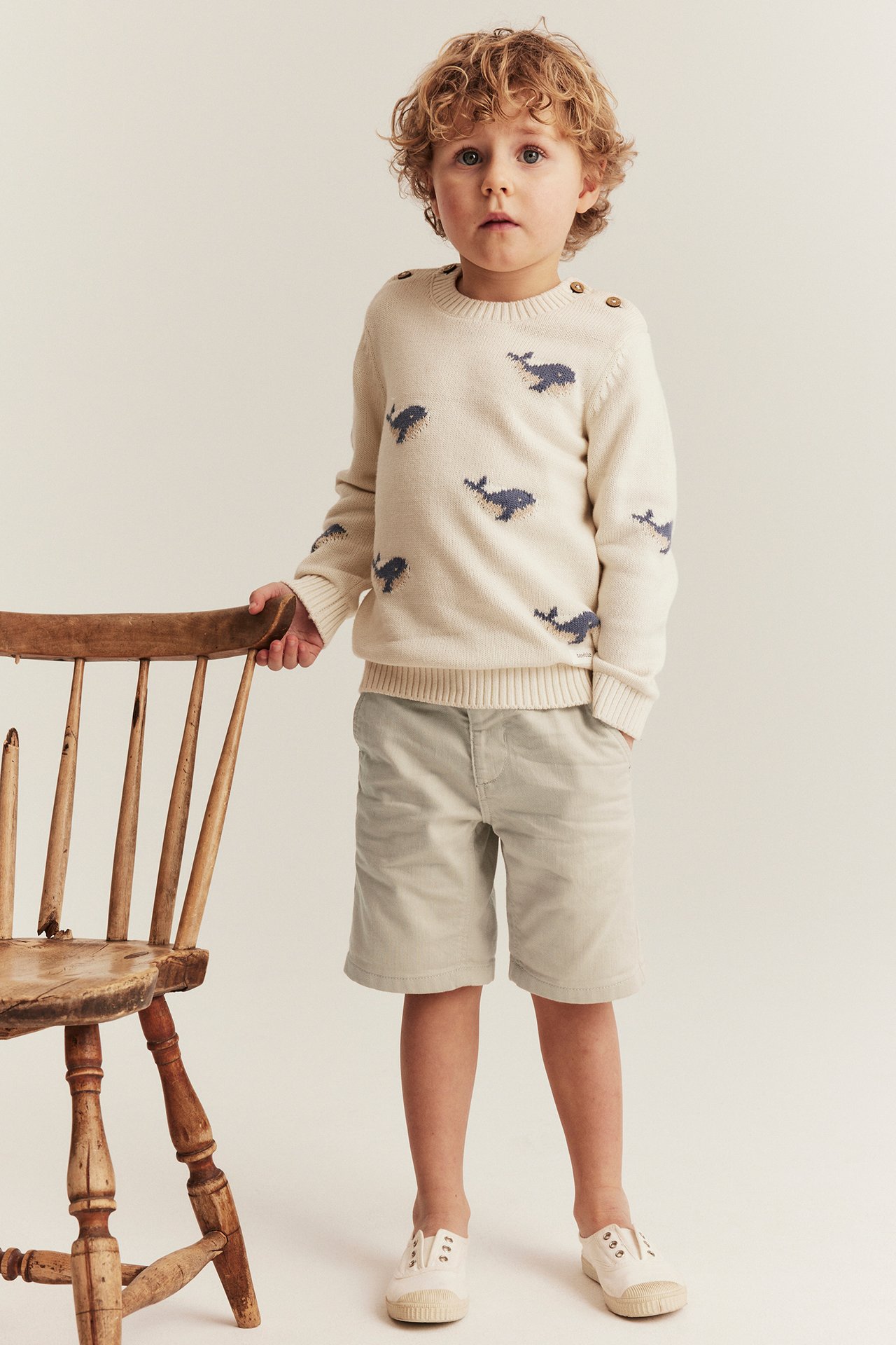 Forfra, et barn har på seg en kremfarget genser med hvalmønster og skulderknapper, samt lysebeige shorts.