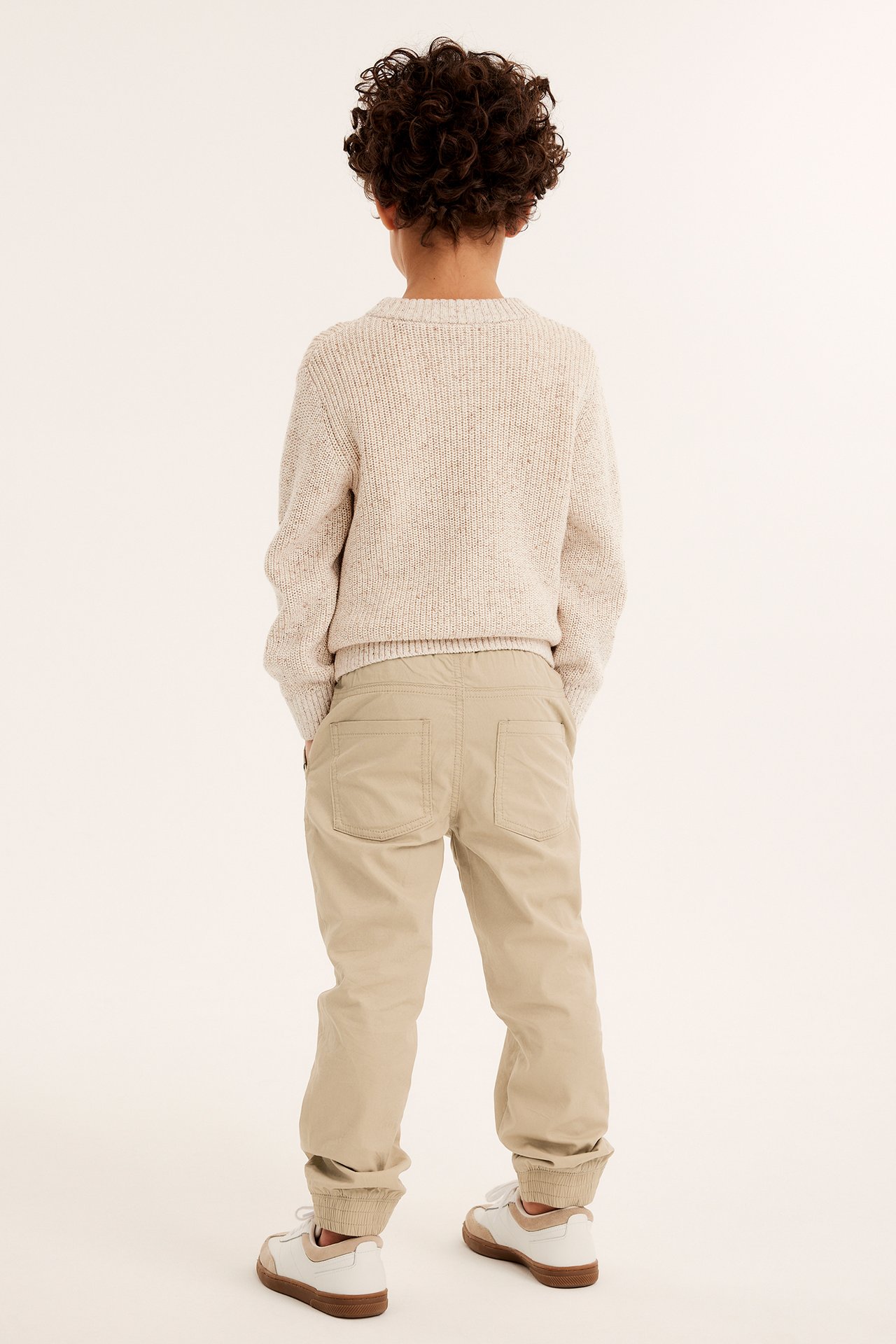 Bakifrån, ett barn i beiga vävda byxor med resår, en ljusbeige stickad tröja och vita sneakers.