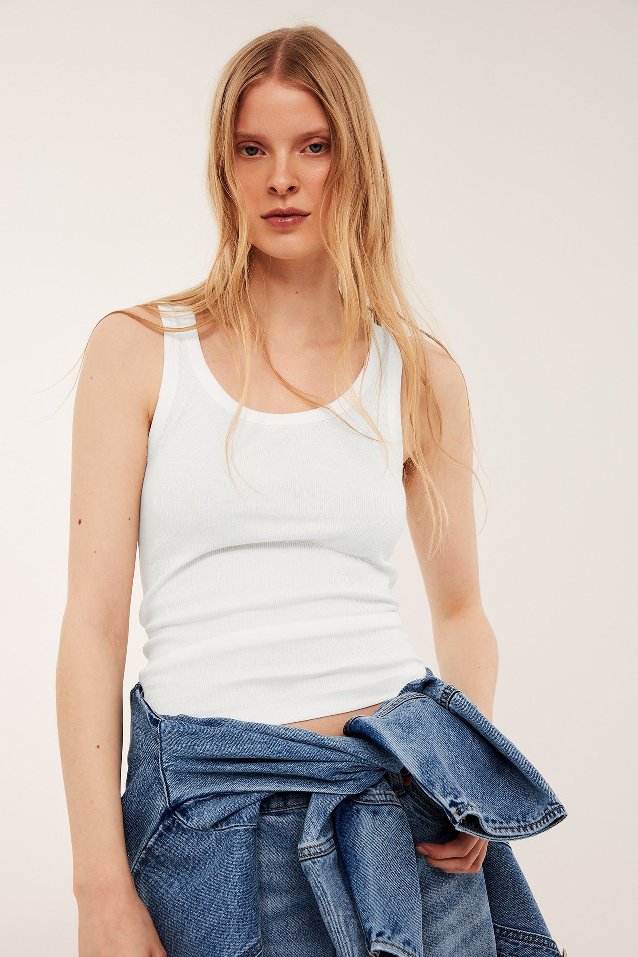 Frontvy av en kvinna i ett offwhite ribbat linne, med en blå jeansjacka knuten runt midjan.