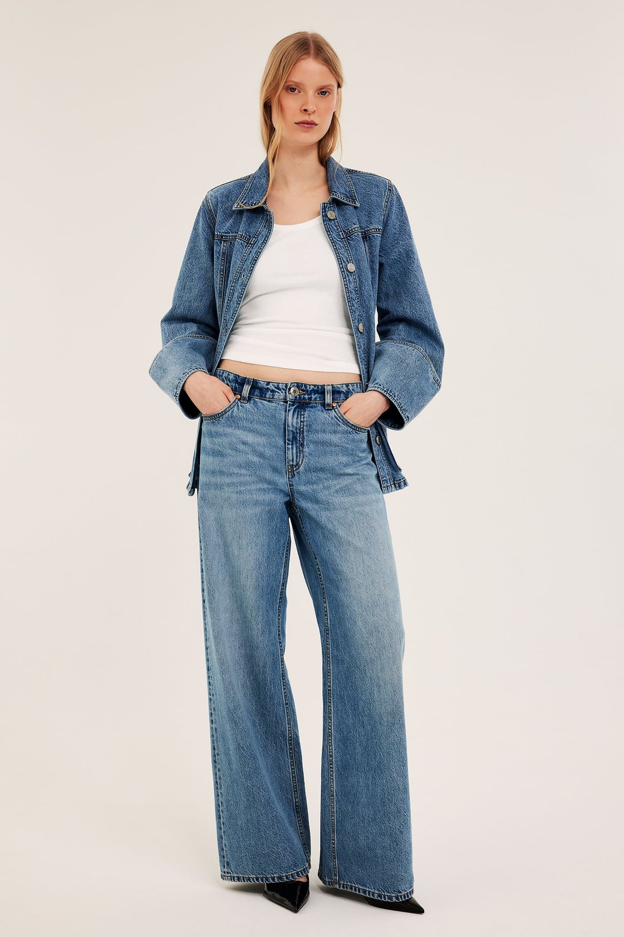 Forfra, kvinne i offwhite ribbestrikket singlet, blå dongerijakke, vide jeans, hendene i lommene.