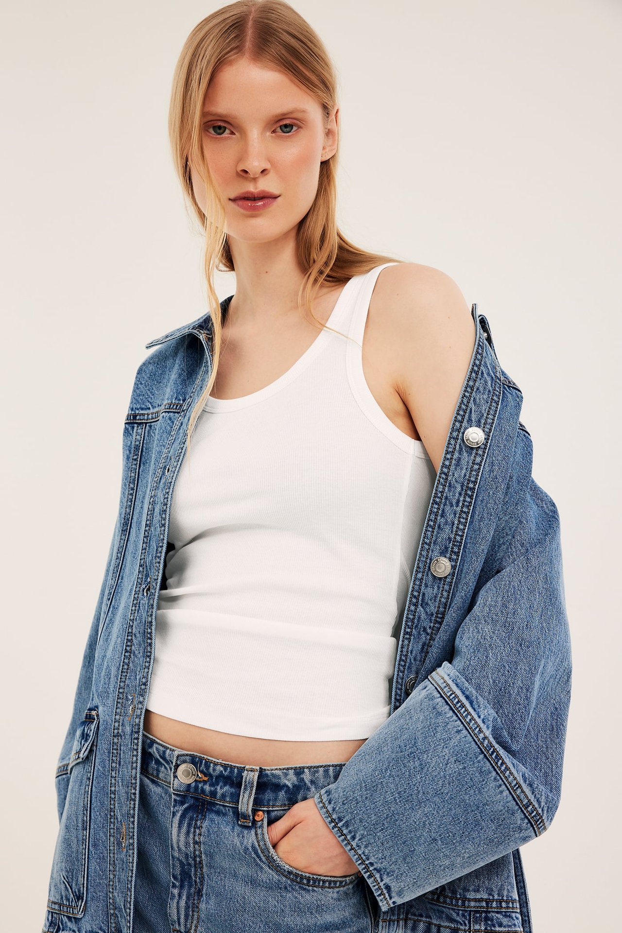 Frontbilde av kvinne i hvit ribbestrikket singlet, blå dongerijakke og jeans med hånd i lommen.