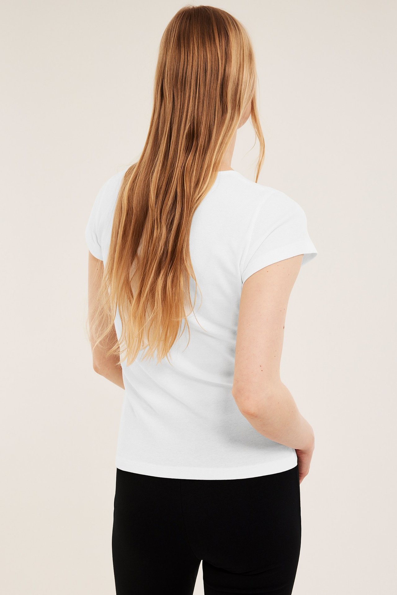 Bakfra av en kvinne i en tettsittende offwhite ribbestrikket kortermet t-skjorte og svarte bukser.