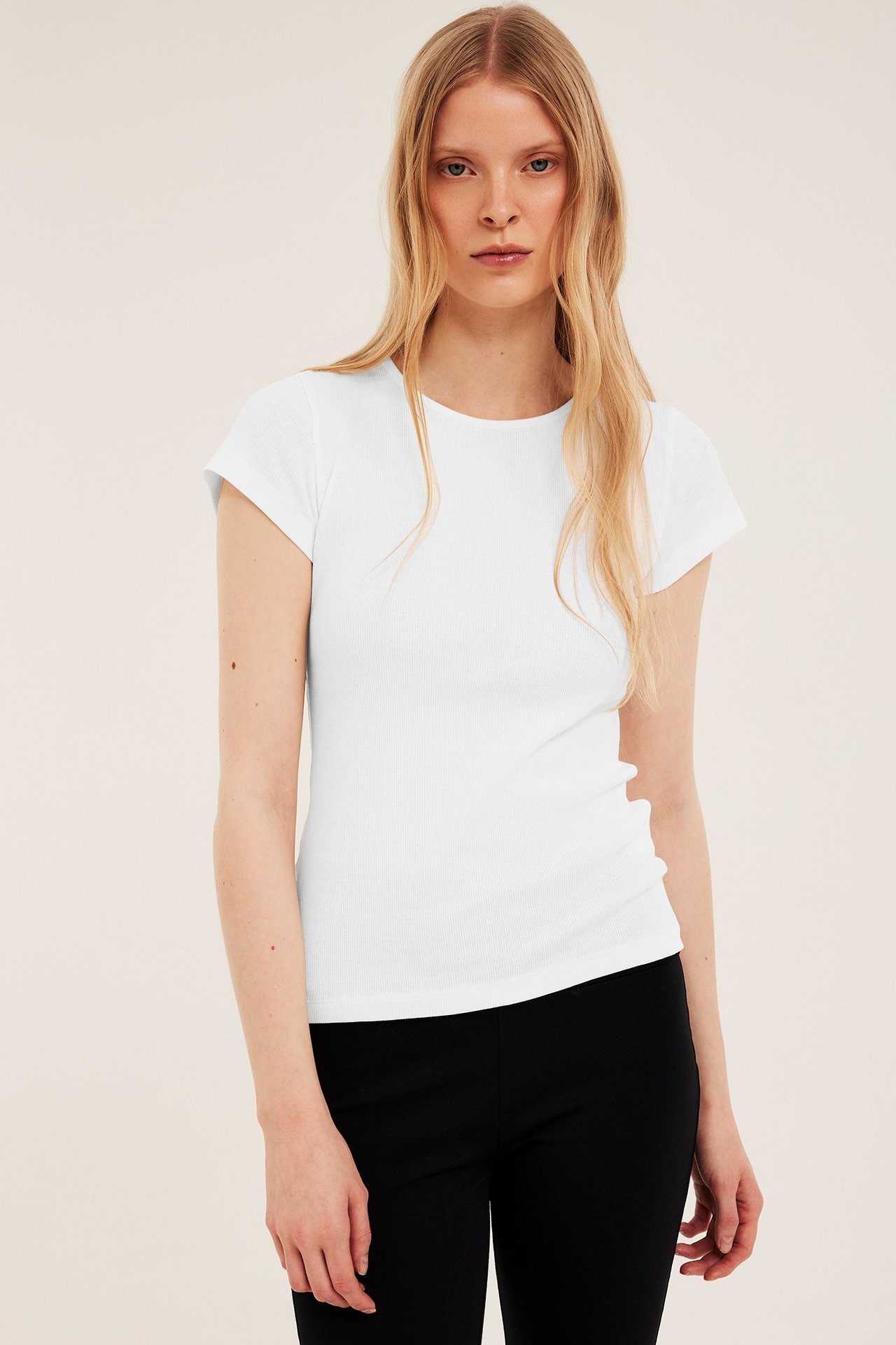 Forfra: Kvinne i figurnær offwhite ribbestrikket T-skjorte med korte ermer og svarte bukser.
