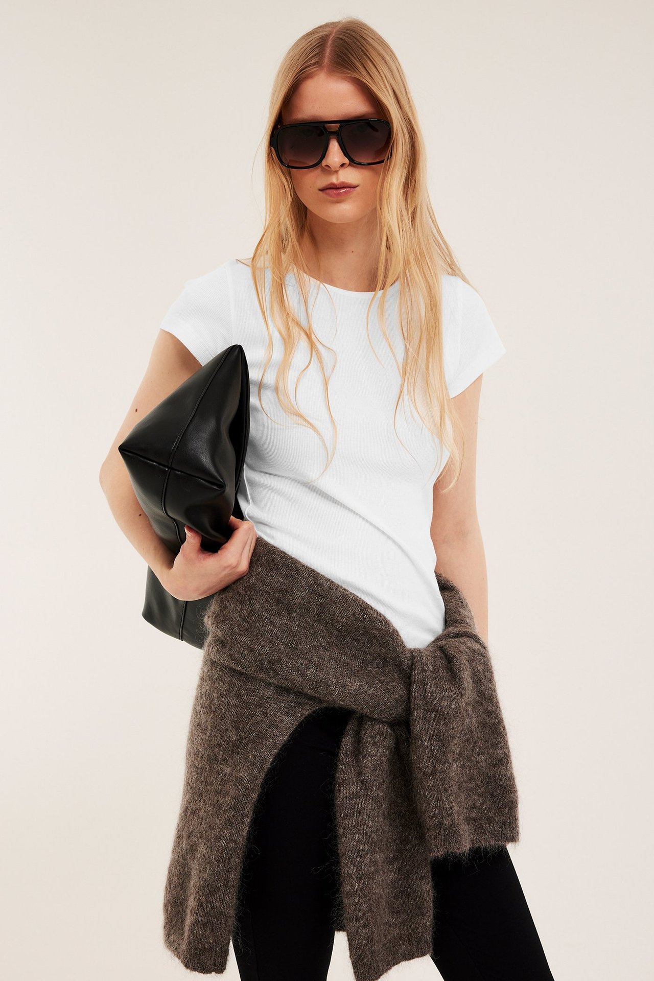Frontbilde: Kvinne i offwhite ribbet t-skjorte, svarte bukser, brun genser, solbriller, holder sort veske.