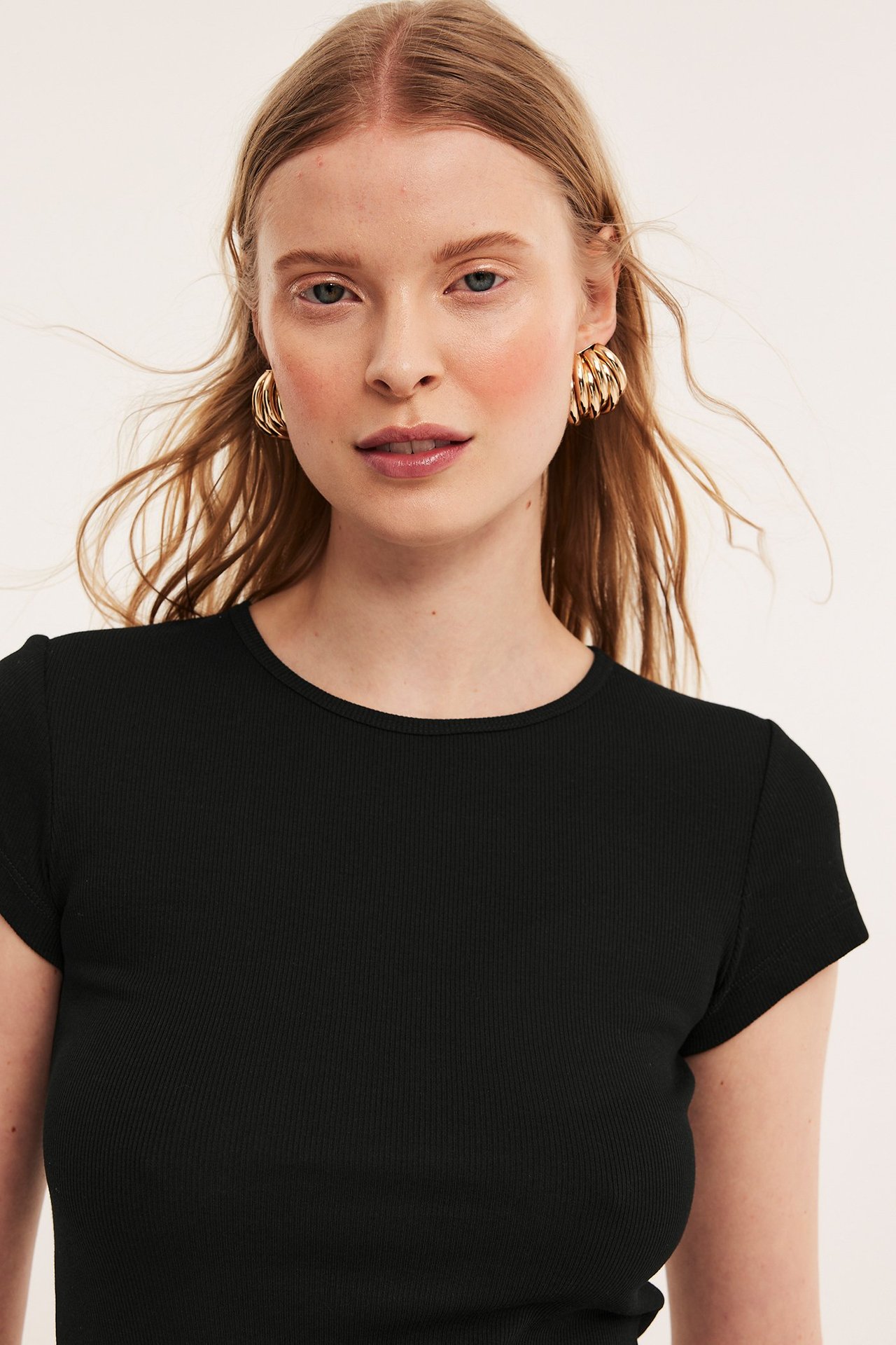 Framifrån, kvinna i svart kortärmad åtsittande ribbad t-shirt och guldiga ringörhängen.
