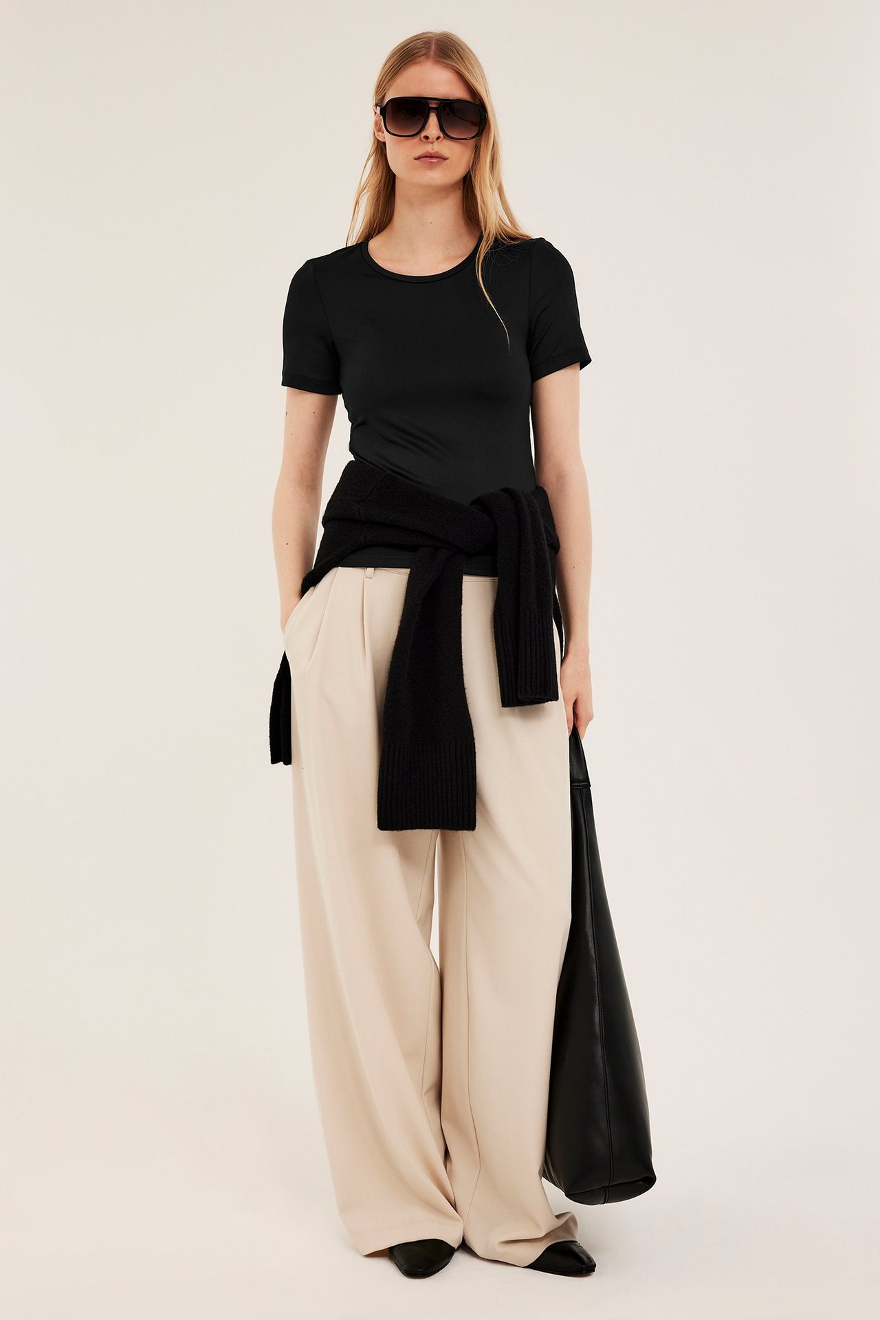 Framifrån: kvinna i svart ribbad t-shirt, beiga vida byxor, svart tröja, solglasögon och svart tygväska.