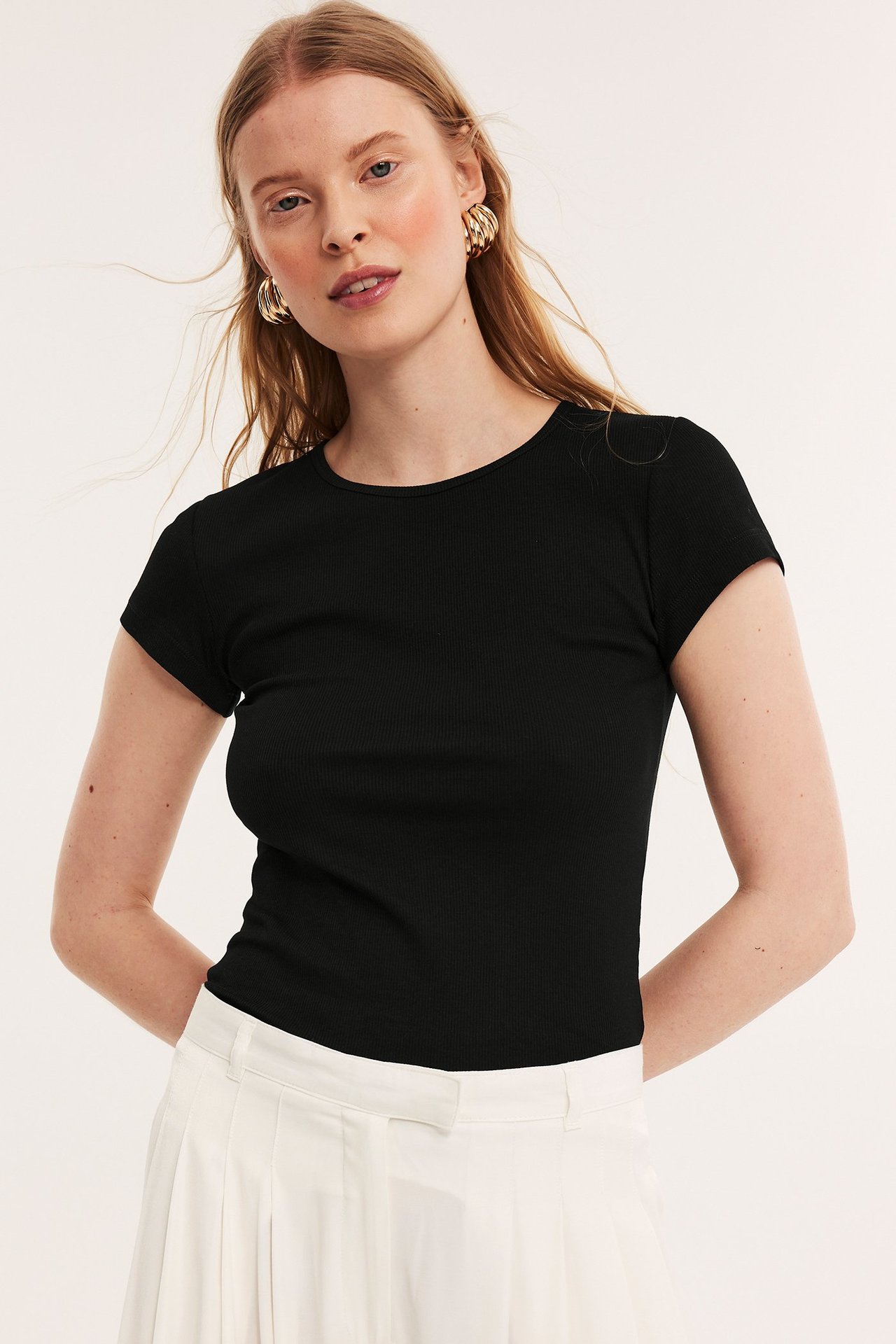 Frontvy av kvinna i svart ribbad kortärmad t-shirt, vit plisserad kjol och guldiga ringörhängen.