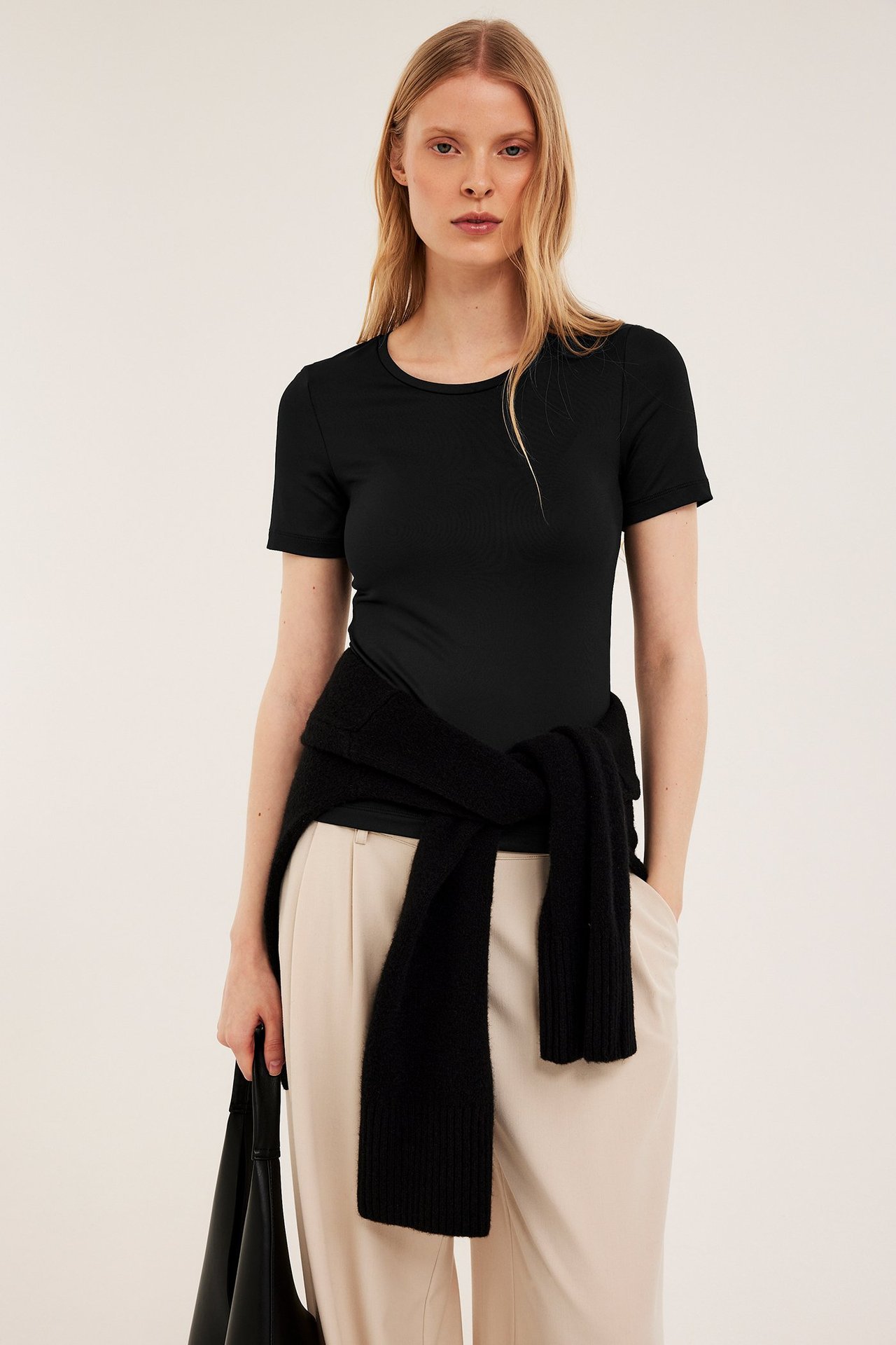 Widok z przodu: kobieta w czarnym prążkowanym t-shircie, beżowych spodniach, czarny sweter w talii.