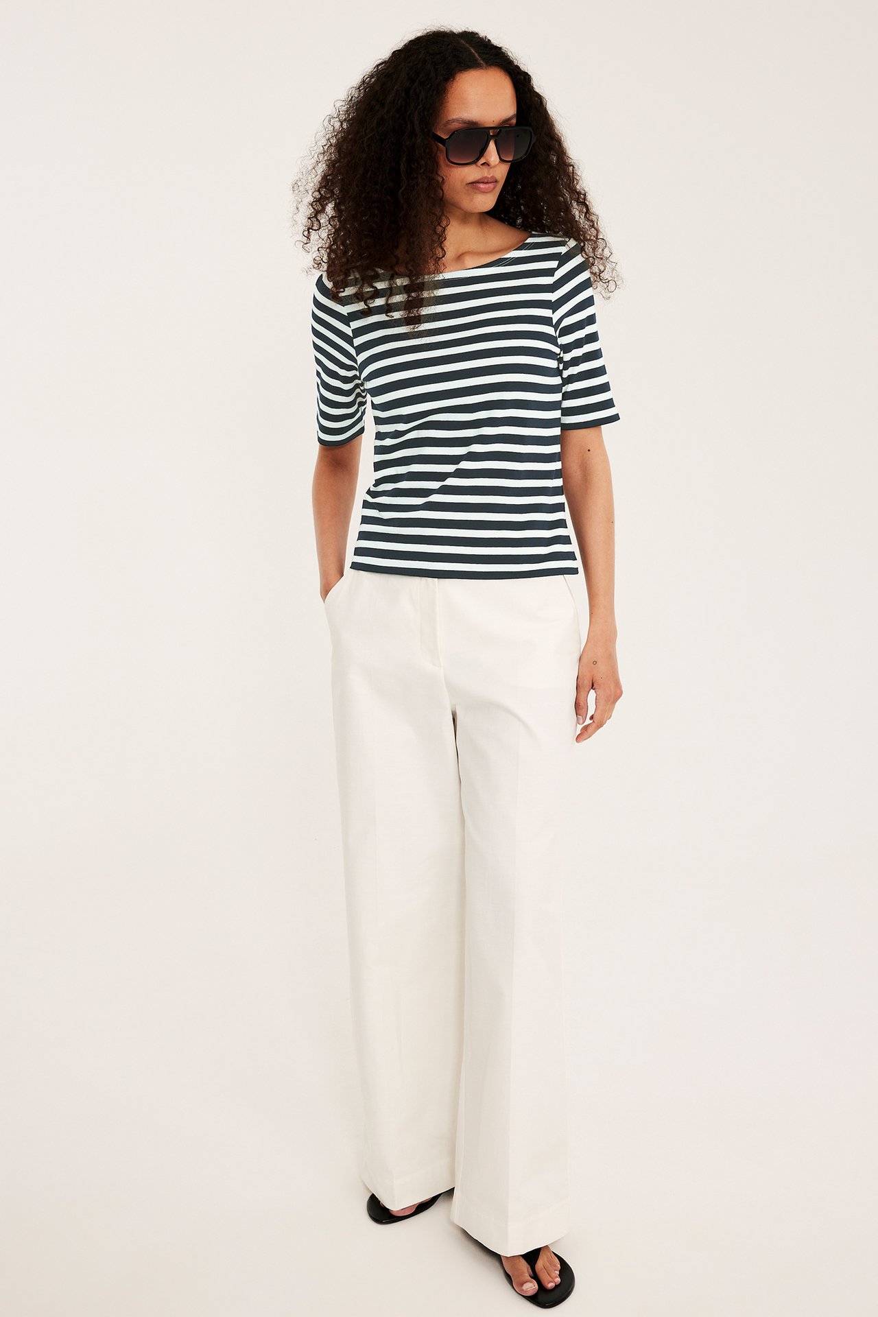 Edestä: Nainen yllään tummansininen ja luonnonvalkoinen raidallinen venepääntiepusero, luonnonvalkoiset leveät housut, mustat sandaalit.