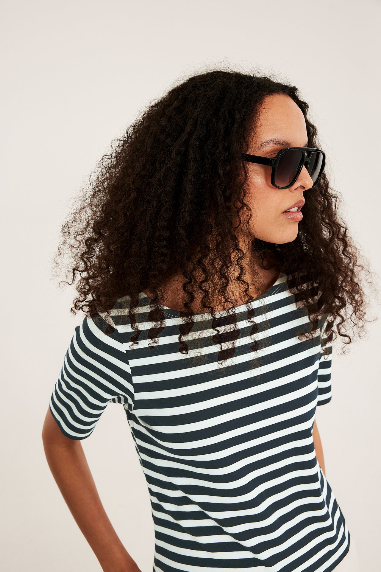 Nainen raidallisessa offwhite lyhythihaisessa venekaula-aukkoisessa topissa ja mustissa aurinkolaseissa, sivukuva.